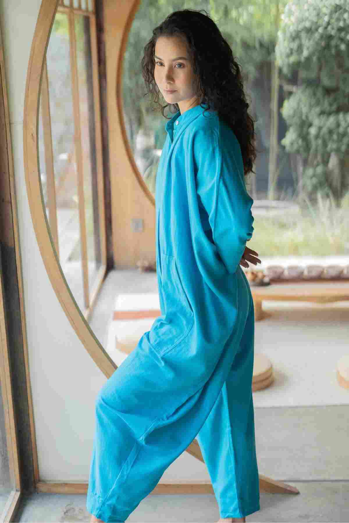 A person with curly dark hair stands indoors by a large circular window, wearing a loose-fitting, limited edition bright blue Miracle Playsuit 2.0 from the Karama Collection by Myrah Penaloza. They are looking toward the camera with a calm expression, their hands behind their back. The background features greenery outside the window, adding to the boho chic vibe.