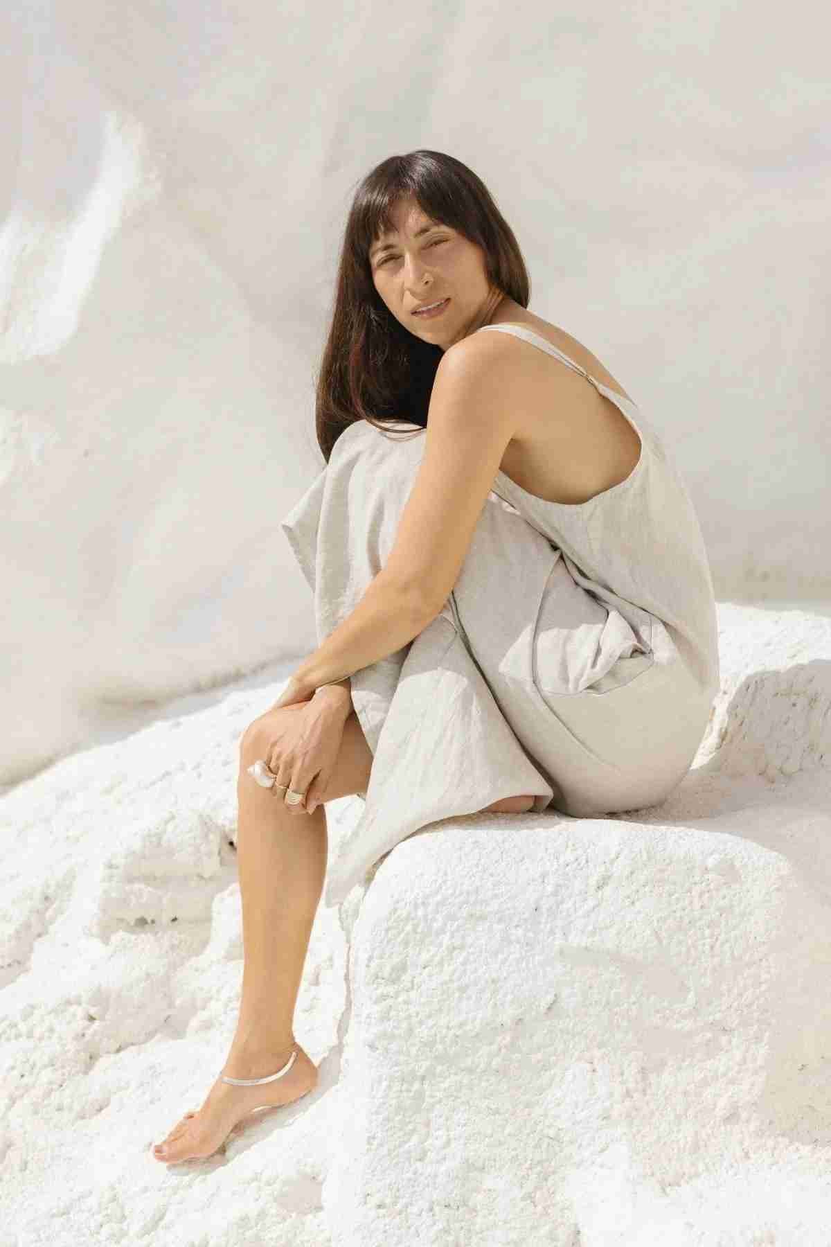 A person with long dark hair, wearing a light beige *Everyday Linen Slip* from Myrah Penaloza and white sandals, is sitting on a white rock formation. The background is also a bright white, creating a clean and serene atmosphere. The person appears relaxed and content, enjoying the luxurious comfort of the slip dress and gazing slightly off to the side.