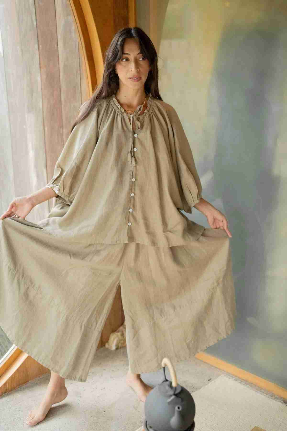 A woman is standing in front of a wooden and glass wall, dressed in a loose-fitting, beige ensemble featuring the Pantalon Linen Pants (Wide Legged) by Myrah Penaloza. She gently holds out the sides of her pants while smiling softly. The warm, natural lighting accentuates the oversized fit of her outfit.