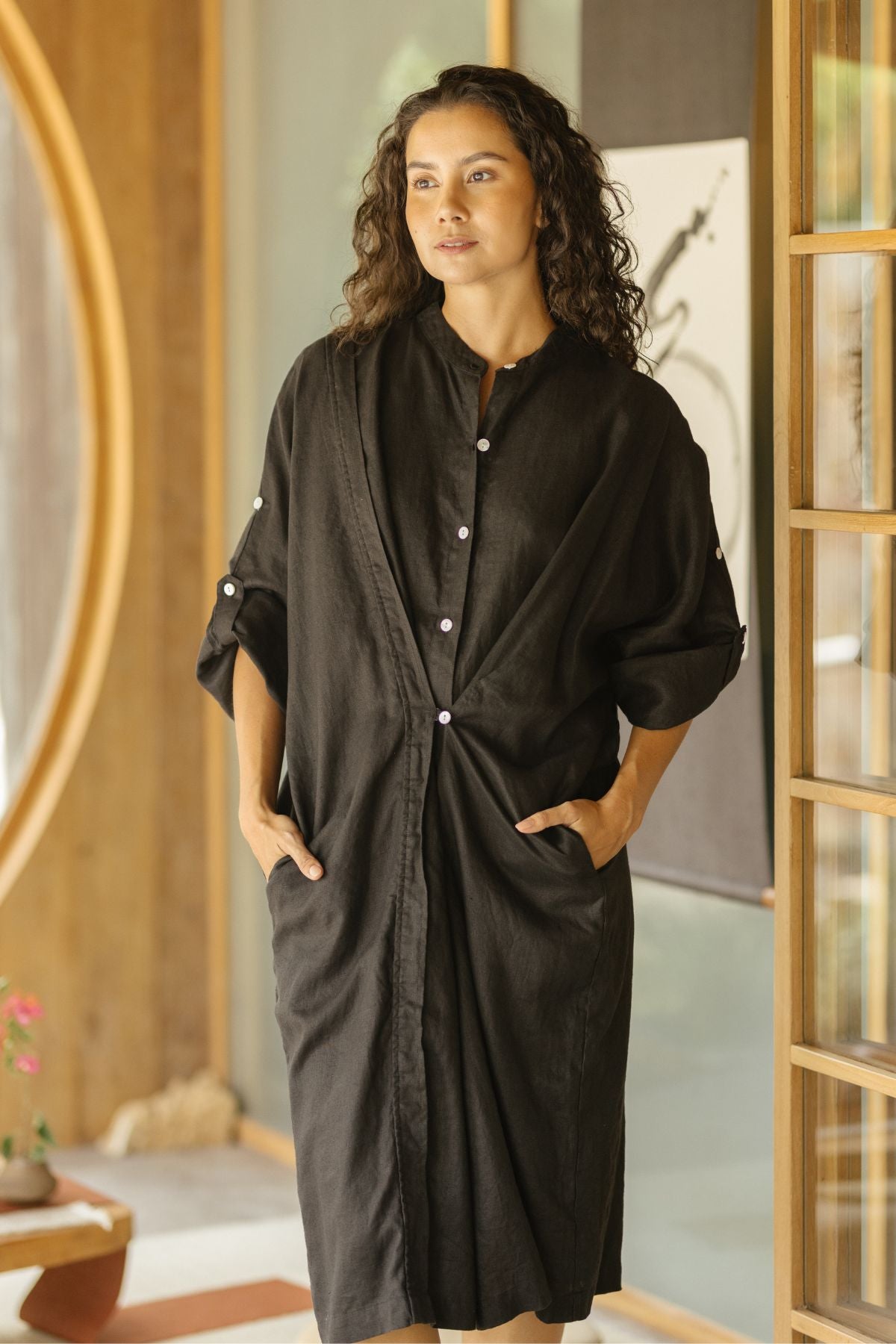 A person with long, curly hair is standing indoors, wearing the versatile Myrah Penaloza Linen Still Water Tunic (Made to Order), a loose-fitting black dress made from 100% linen with rolled-up sleeves and hands in pockets. In the background, there is a circular window and partially visible wall art.