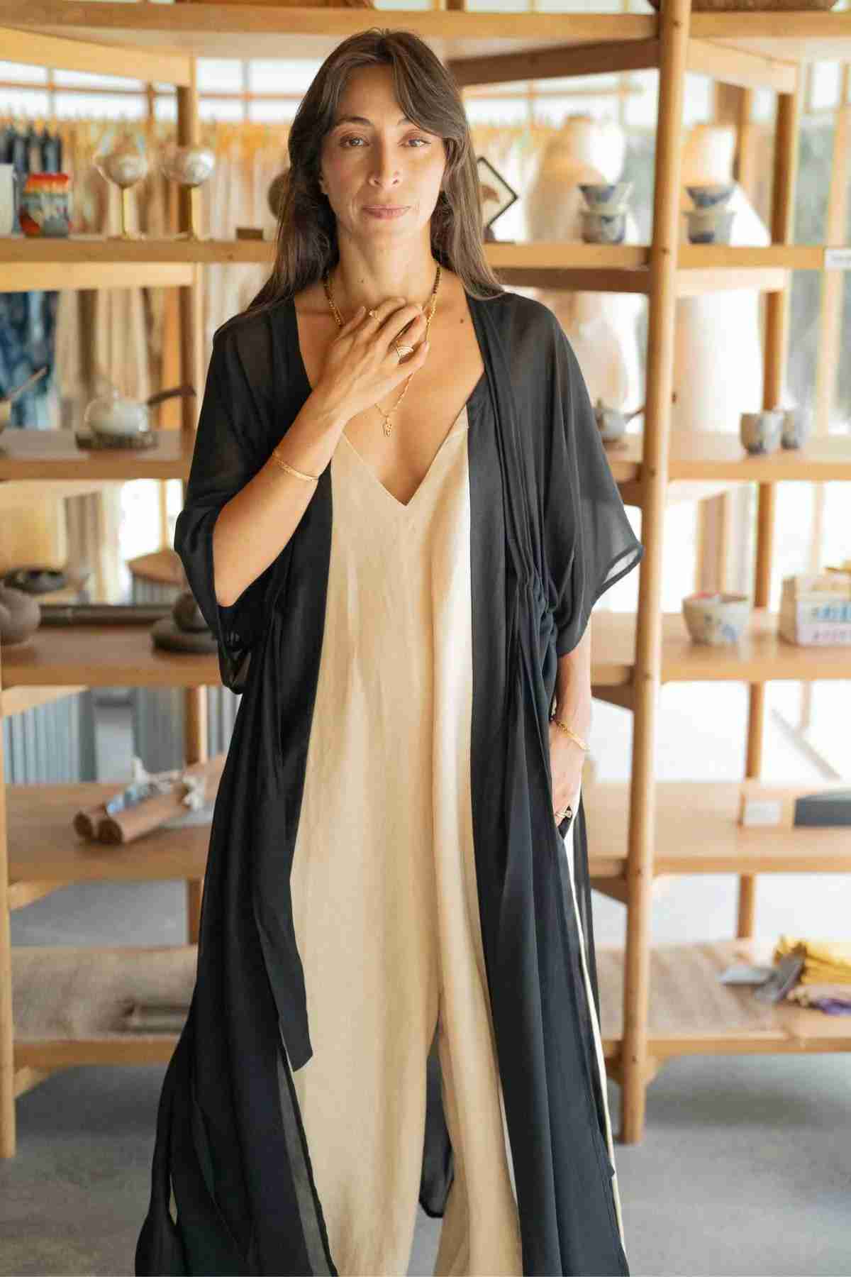 A woman with long dark hair stands in a store with wooden shelves filled with various items. She is wearing a cream-colored dress paired with a Myrah Penaloza Madelin Sheer Chiffon Long Cover, and she has her hand resting gently near her neck. The background showcases an assortment of ceramics and textiles, highlighting sheer brilliance.