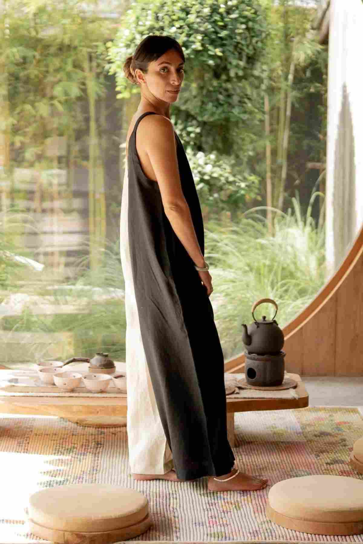 A woman stands barefoot on a patterned rug in a minimalistic, serene tea room. She is dressed in the luxuriously soft Kundalini Linen Playsuit with wide-leg bottoms by Myrah Penaloza. Behind her is a wooden table with tea sets and a black teapot. The background features lush greenery visible through a large window.