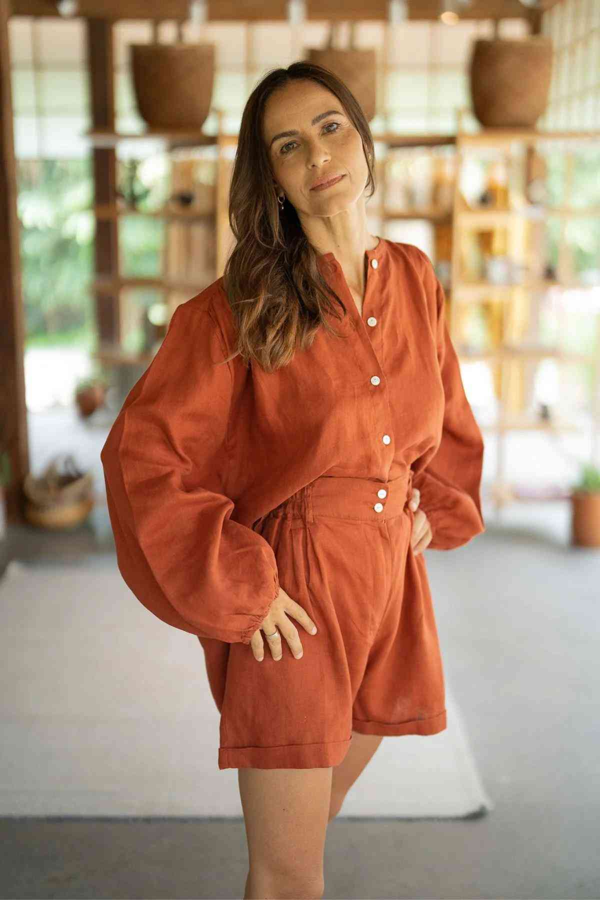A woman in an orange Rice Linen Set from Myrah Penaloza stands indoors with a confident expression. She has one hand on her hip and is in a room with wooden shelves and potted plants, giving the space a cozy, natural feel—showcasing the versatile fashion of elegant stone-washed linen attire.