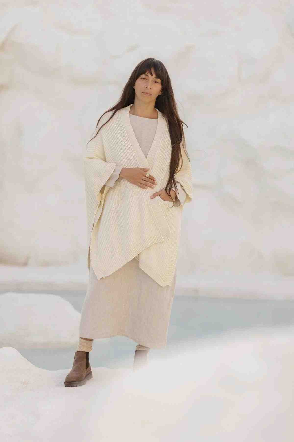A person with long hair stands outdoors against a light-colored background, wearing the Mountain Magic Linen Gown by Myrah Penaloza. Draped over the cream-colored, long knitted shawl paired with their light beige dress and brown boots, they touch the sides of the shawl and look towards the camera, effortlessly embodying elegance in this versatile made-to-order gown.