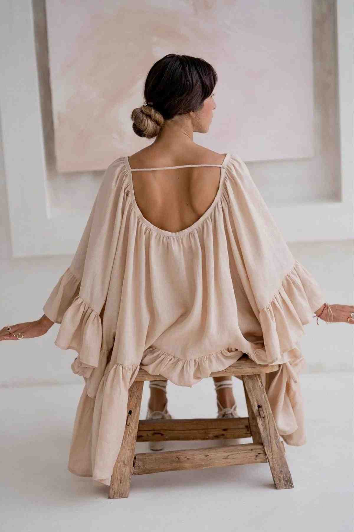 A woman sits on a wooden stool wearing the Linen Brida Gown Short from Myrah Penaloza, which features a romantic spring/summer design with flowy beige linen, an open back, and ruffled sleeves. Her hair is styled in a low bun, complementing the soft and neutral-toned background.
