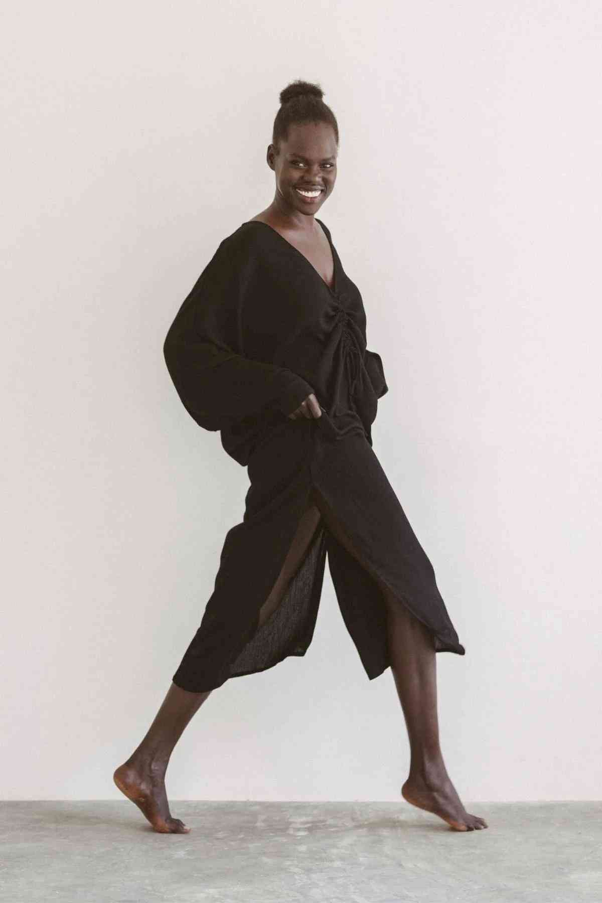 A person with dark skin and a high bun hairstyle is joyfully striding forward. They are wearing a long, black Bamboo Rayon Virgo Rising Gown from Myrah Penaloza, featuring a side slit and have their hands in the pockets. The background is plain and light-colored.