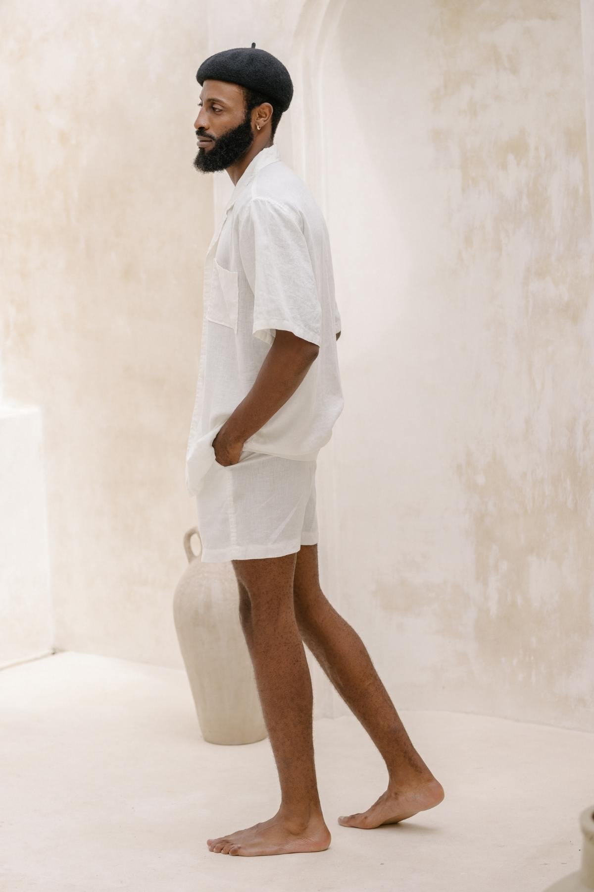 A man wearing a Caravana Men's Linen Top & Short by Myrah Penaloza and a black beret stands barefoot in a minimalist room with a light-colored textured wall. A large ceramic vase is in the background, complementing the serene ambiance, reminiscent of a nomadic traveler’s tranquil retreat.
