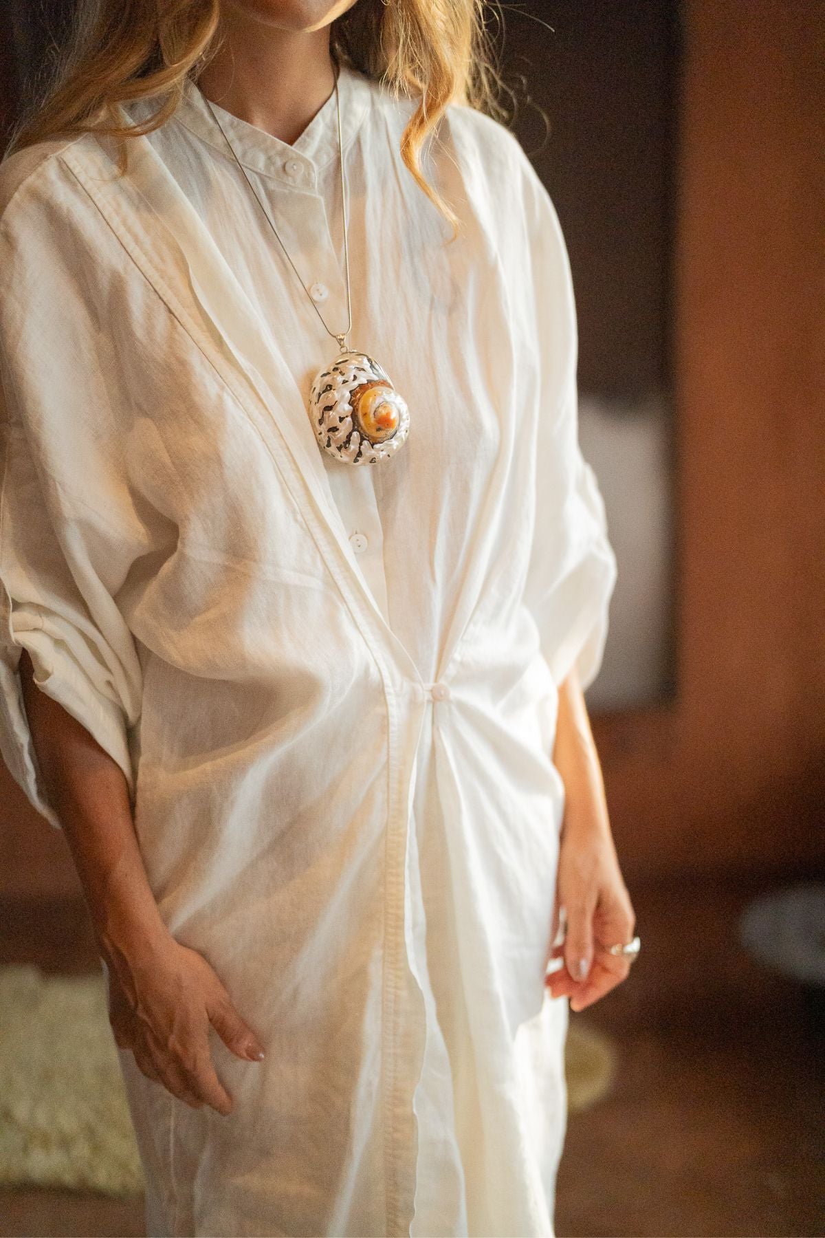 A person wearing a loose, white, long-sleeved Linen Still Water Tunic from Myrah Penaloza, adorned with a large pendant necklace featuring a unique design. The setting appears to be indoors with warm lighting and a soft, blurred background. Only the upper body and part of the arms are visible.