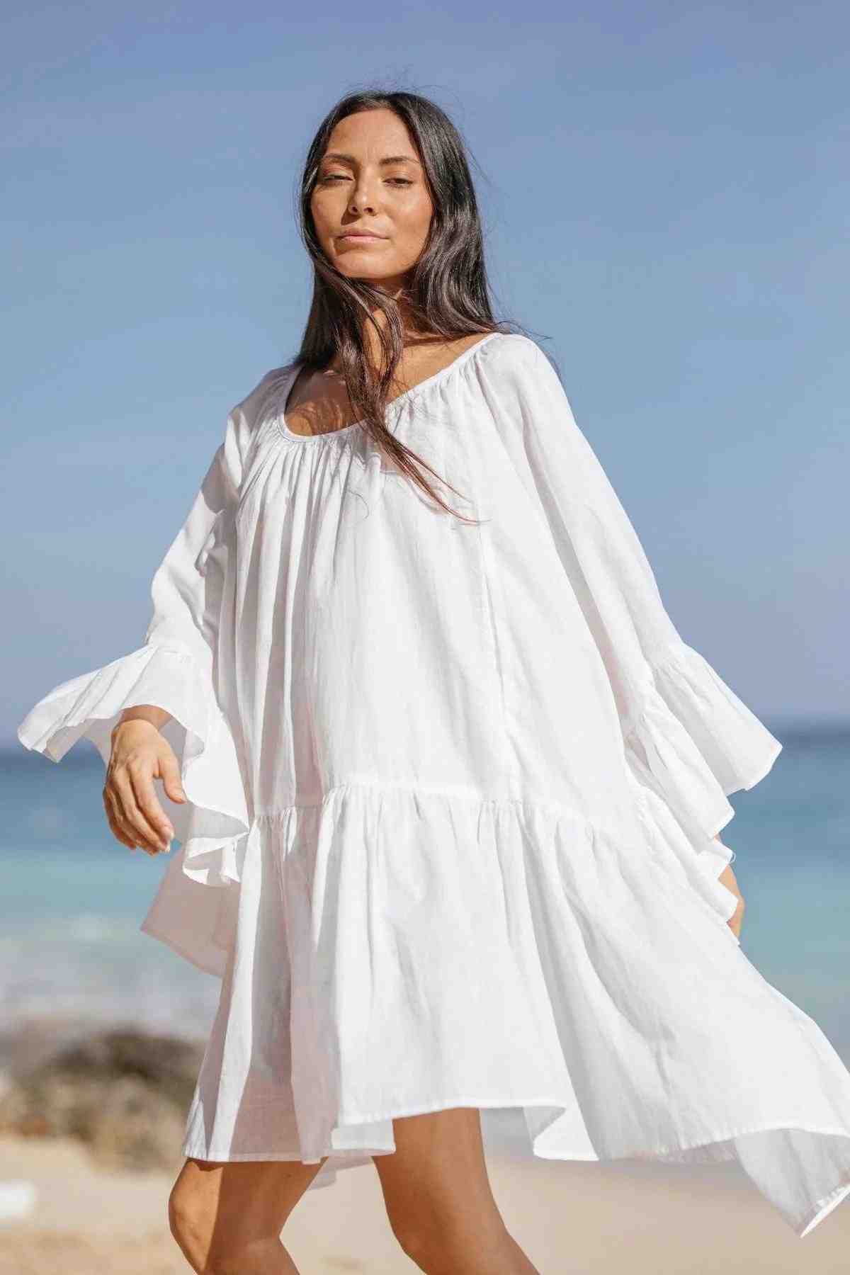 A woman with long dark hair wears the Linen Brida Gown Short, a romantic piece by Myrah Penaloza crafted from linen fabric. She stands on a beach, the airy white gown catching the breeze against the backdrop of a clear blue sky and ocean waves.