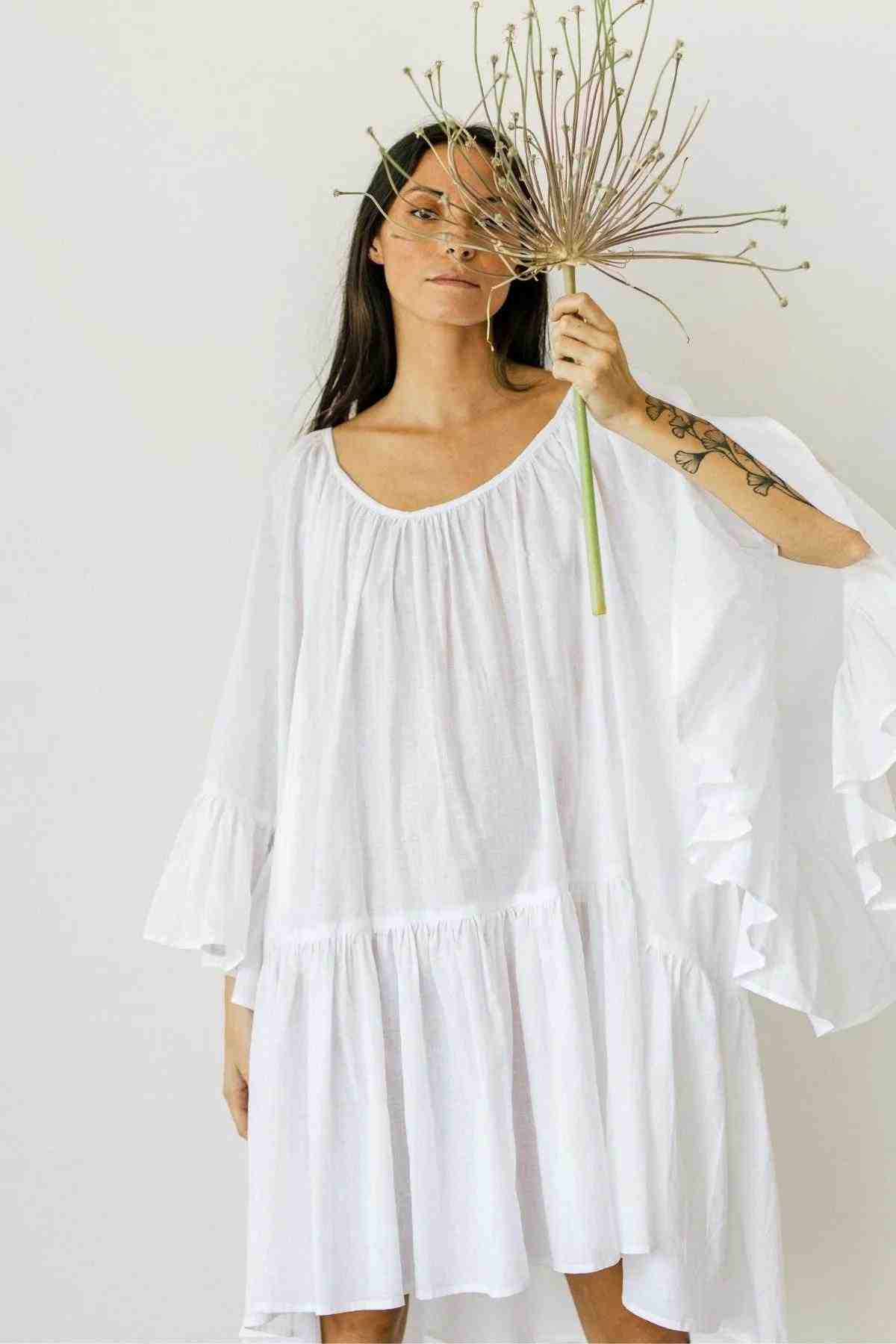 A woman in a flowy Myrah Penaloza Linen Brida Gown Short holds a large, dried allium flower in front of her face. The romantic spring/summer dress contrasts with the plain white background, as her left arm extends to reveal a tattoo on her forearm.