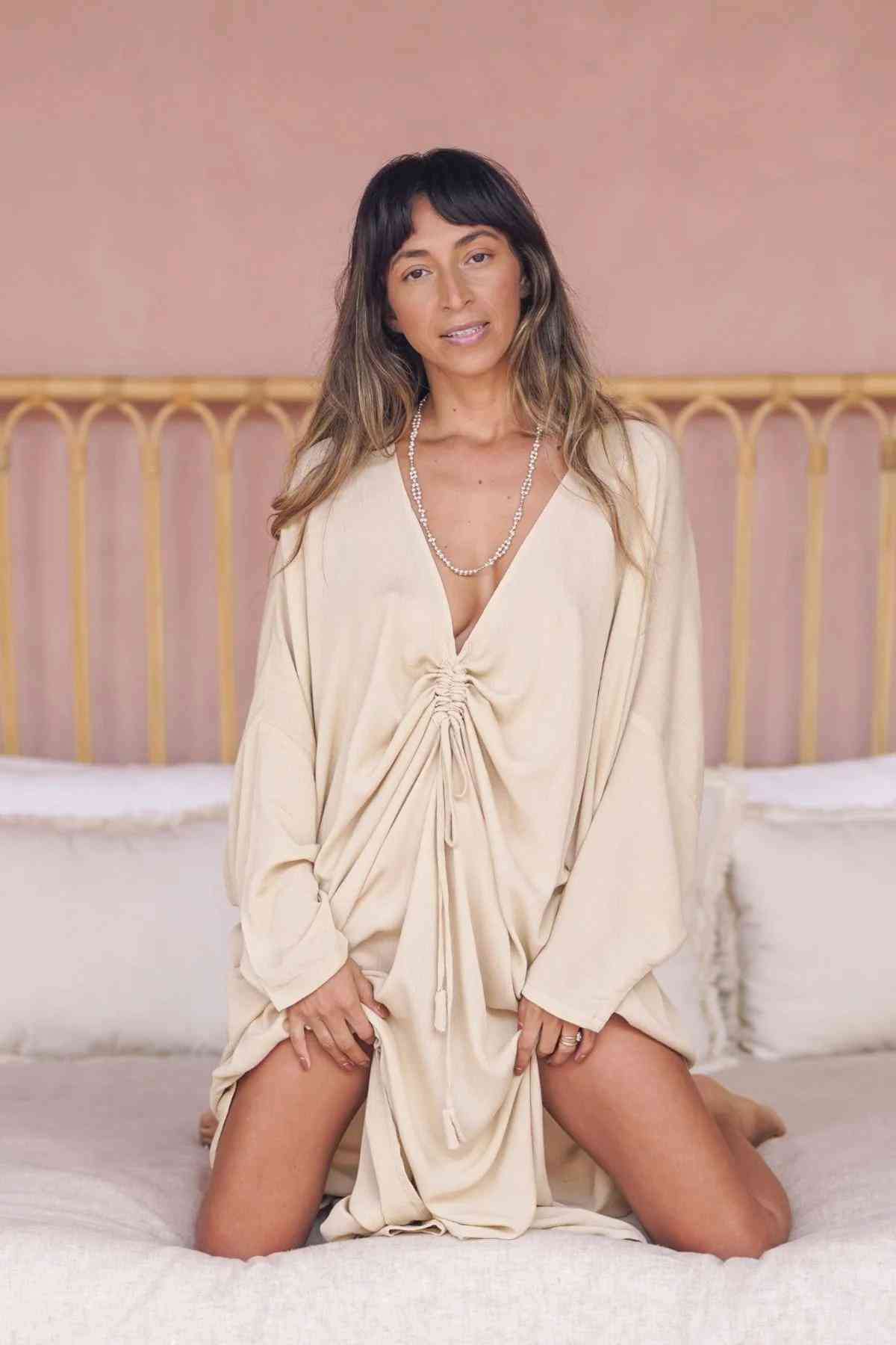 A person with long hair is kneeling on a bed with white linens, wearing a Myrah Penaloza Bamboo Rayon Virgo Rising Gown. This elegant gown features a light color and a drawstring at the front. They are smiling and looking at the camera in a room adorned with warm, soft pink walls and a headboard made of light wood.