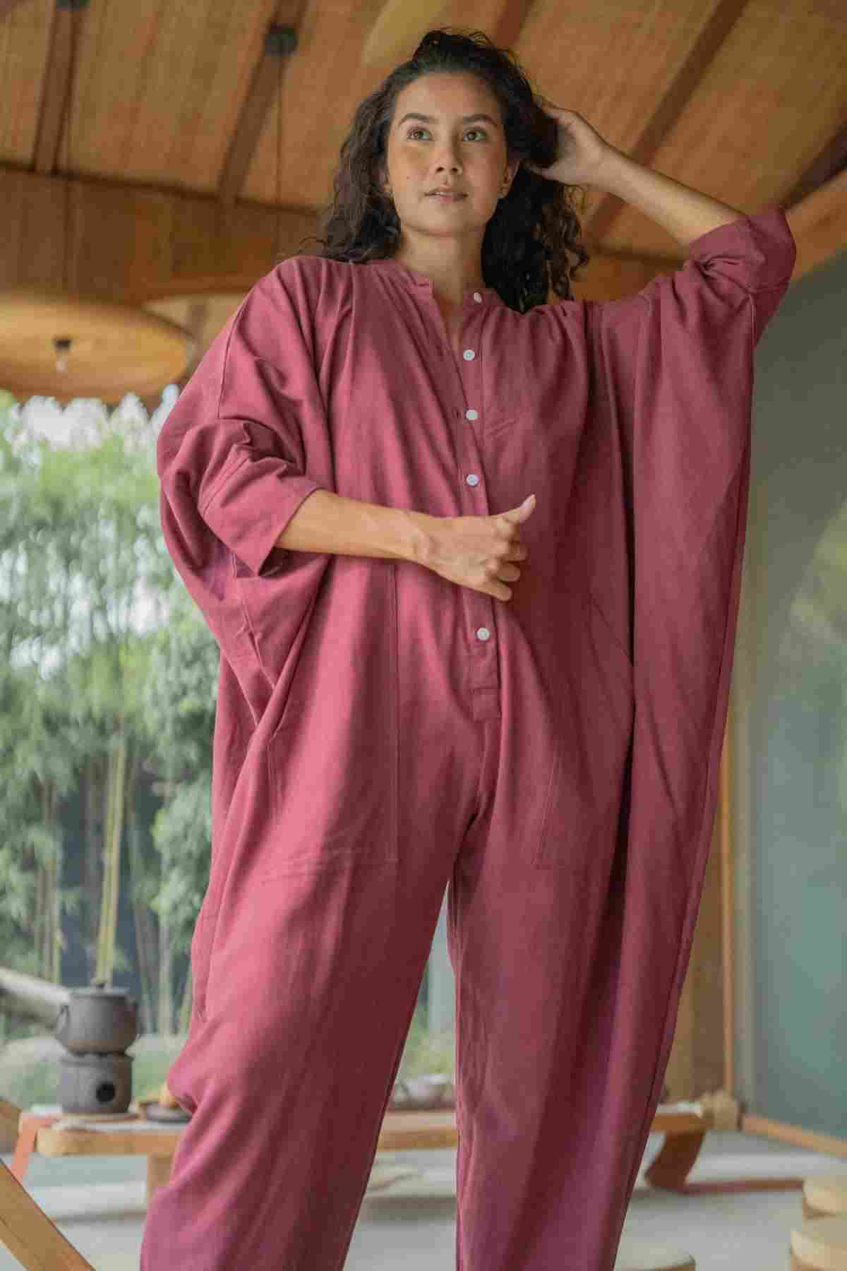 A person stands confidently in a wooden veranda, wearing the limited edition Miracle Playsuit 2.0 from the Karama Collection by Myrah Penaloza, a loose-fitting maroon jumpsuit with buttons down the front. One hand holds their hair back while the other is placed at the waist. The boho chic setting features green foliage and part of a structure with large windows in the background.