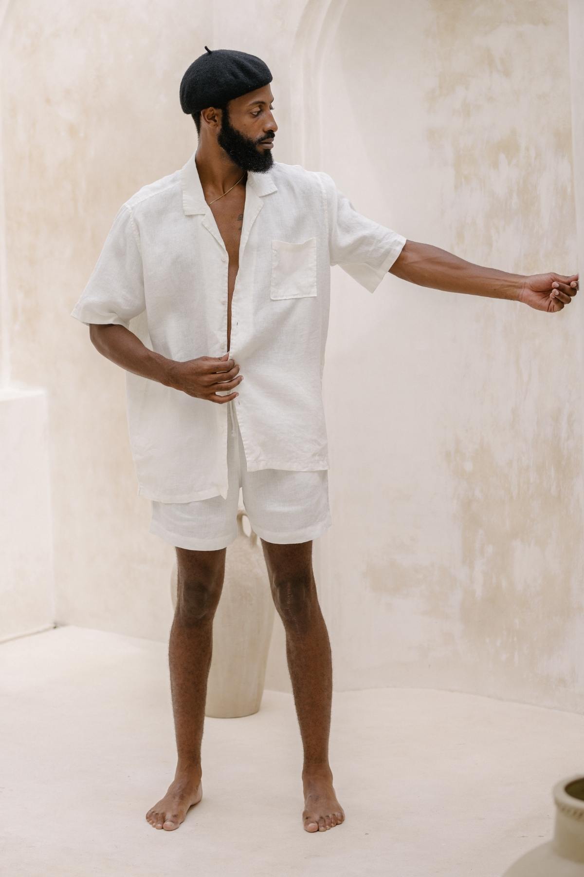 A nomadic traveler with a beard stands in a minimalist setting, wearing the lightweight and breathable Caravana Men's Linen Top & Short by Myrah Penaloza, topped with a black beret. His outstretched arm points forward as he gazes in that direction, set against neutral-toned walls.