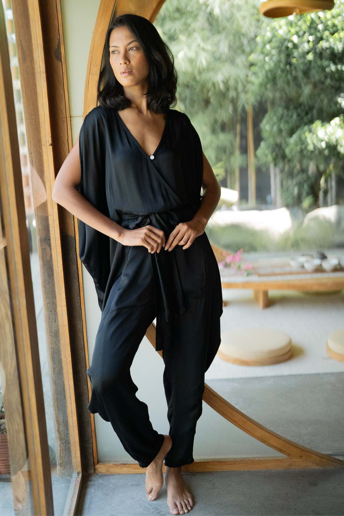 A woman in a Kuan Yin Playsuit of 100% silk, botanically dyed in turmeric gold by Myrah Penaloza, stands barefoot beside a large wooden-framed window. She gazes contemplatively outside at the blurred, lush green garden background. The room exudes minimalist natural decor with French linen accents.