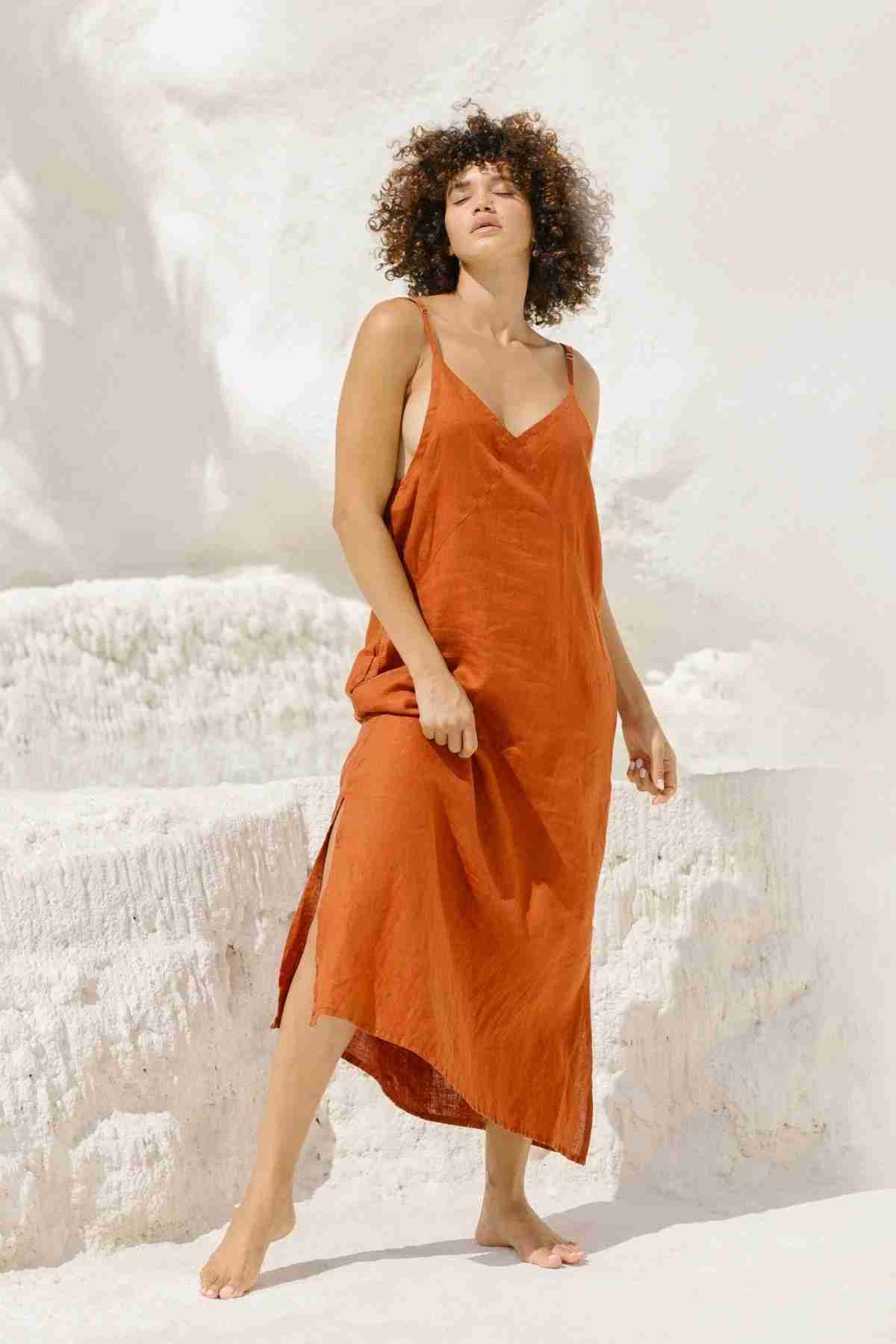 A woman with curly hair stands barefoot on light-colored rocky terrain, wearing a loose, rust-orange sleeveless Everyday Linen Slip by Myrah Penaloza, characterized by its thin straps. She gazes slightly upward with one leg forward and her left hand resting on her dress. The scene is bright and sunlit.