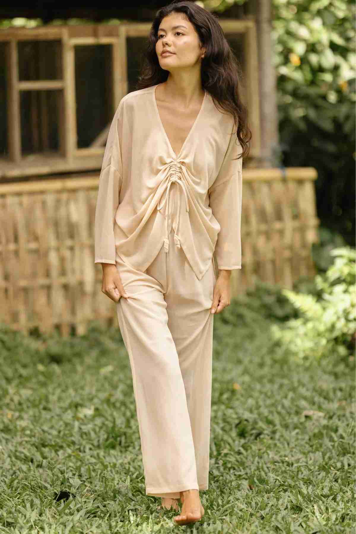 A person with long dark hair stands on a grassy area, adorned in the luxurious Chiffon Silk Sheer Virgo Rising Set from Myrah Penaloza. The ensemble includes a chiffon silk sheer top with a tie detail at the front and matching pants. Behind them, a rustic wooden structure and lush greenery create a picturesque background.
