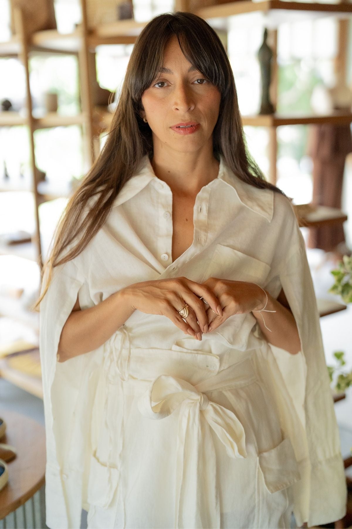 A woman stands indoors, wearing the Sutra Linen Set from Myrah Penaloza. The set features a white blouse with a tied waist and a matching white cape draped elegantly over her shoulders. She has long, straight brown hair and is looking directly at the camera with a neutral expression. Blurred shelves loaded with various items in the background highlight the versatility of her stylish linen ensemble.
