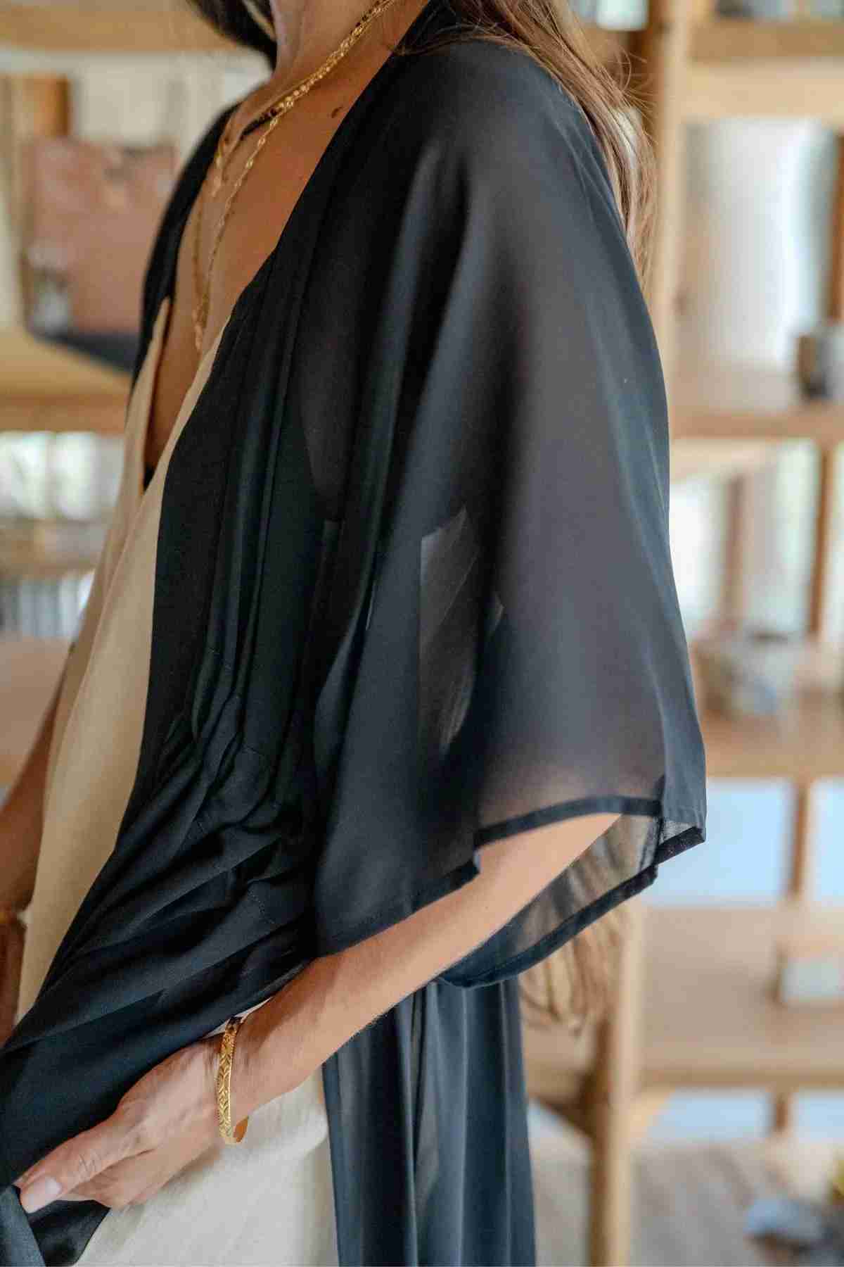 Close-up of a person wearing a Myrah Penaloza Madelin Sheer Chiffon Long Cover over a white outfit. The person is accessorized with gold jewelry, including a necklace and bracelet. The background shows shelves with various items, slightly out of focus.