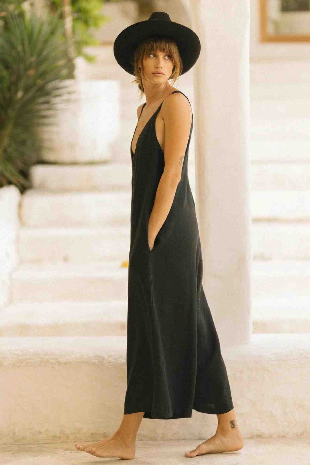 A person standing barefoot on a white, outdoor staircase is wearing the Kundalini Linen Playsuit by Myrah Penaloza. The luxuriously soft stonewashed linen fabric of the playsuit flows elegantly, while a wide-brimmed black hat and shoulder-length hair frame their face. Green plants are visible in the background.
