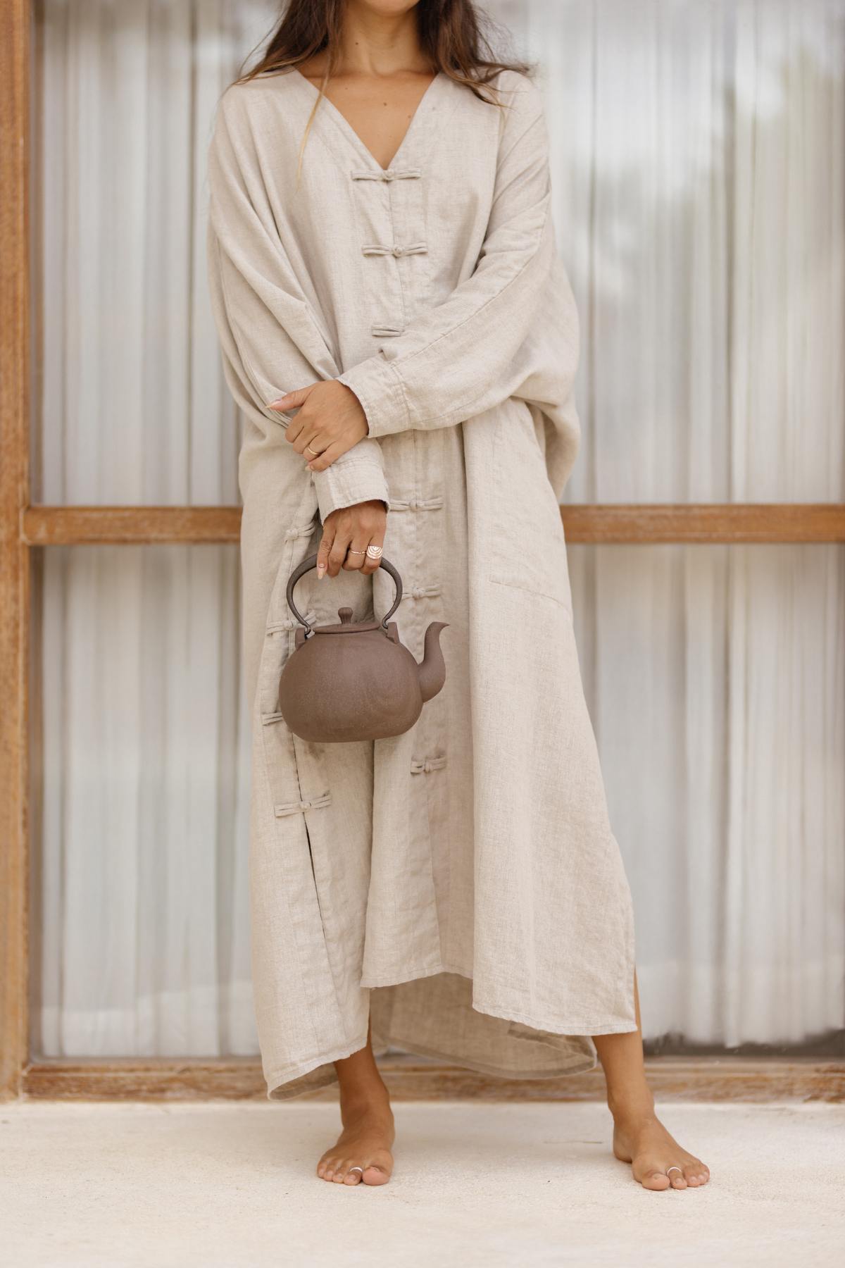 A person stands in front of a window with white curtains, wearing a long, beige Ancient Chajin Jacket (100% Linen) from Myrah Penaloza and holding a brown teapot with a curved handle.