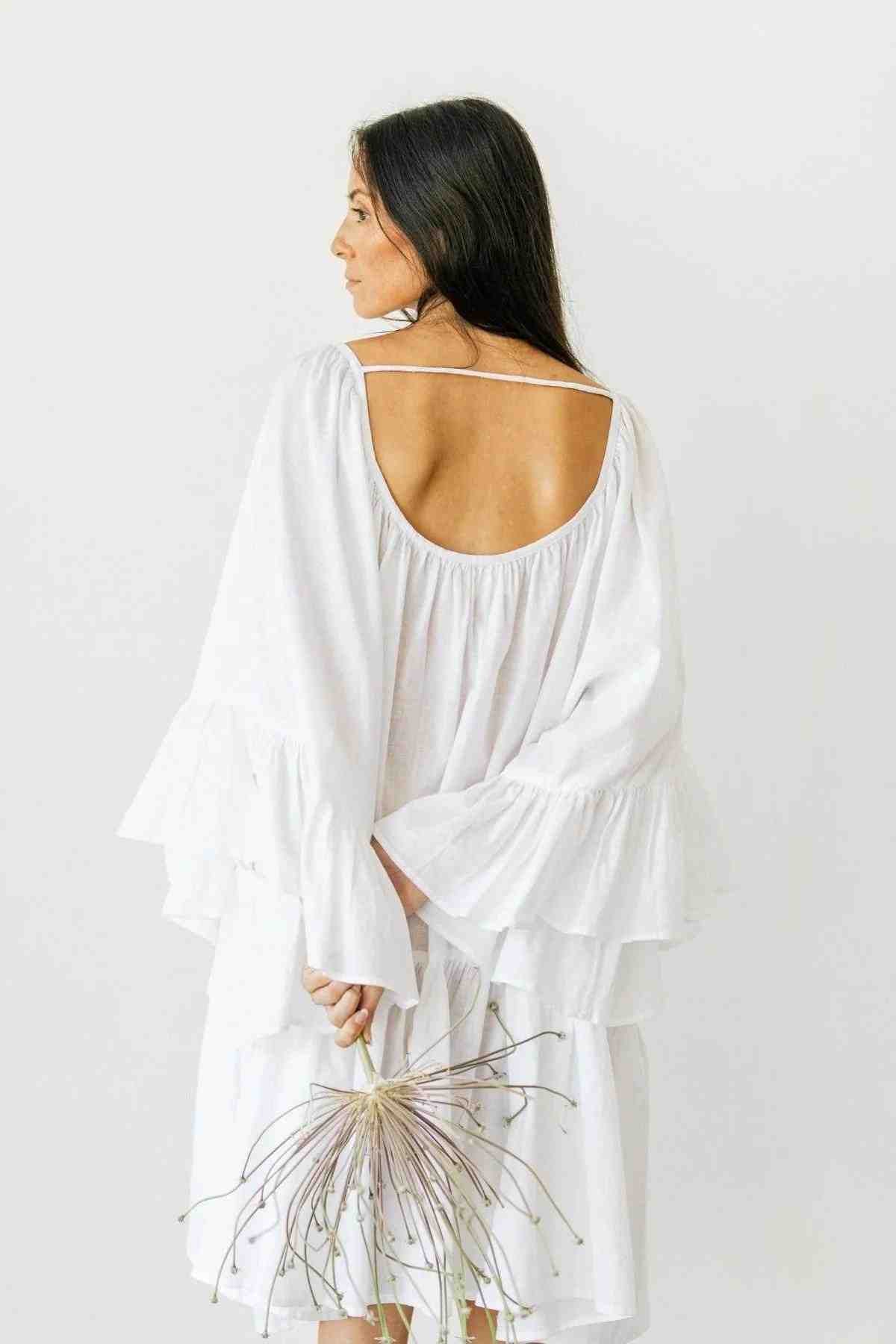 Against a plain backdrop, a woman with long dark hair wears a loose-fitting Linen Brida Gown Short from Myrah Penaloza. The white, ruffled linen dress, featuring an open back, captures the allure of a romantic spring/summer ensemble. She gazes over her shoulder while holding a dried, spiky plant behind her back.