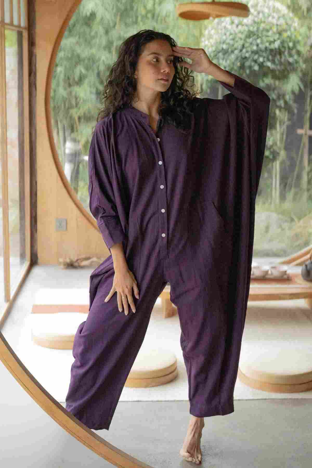 A woman stands barefoot in front of a circular window, wearing the limited edition Miracle Playsuit 2.0 (Karama Collection) by Myrah Penaloza—a loose-fitting, long-sleeved purple jumpsuit with buttons down the front. She has curly hair and strikes a thoughtful pose with one hand on her forehead. The background showcases a modern wooden interior with greenery visible outside.