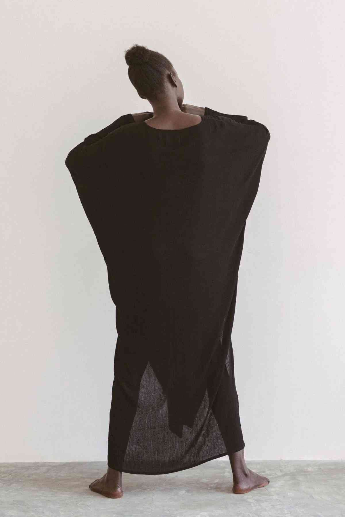 A person stands barefoot against a light background, facing away from the camera. They have dark skin and are wearing a Virgo Rising Gown made of Bamboo Rayon by Myrah Penaloza, which covers their body and extends to the ground, creating a dramatic silhouette.