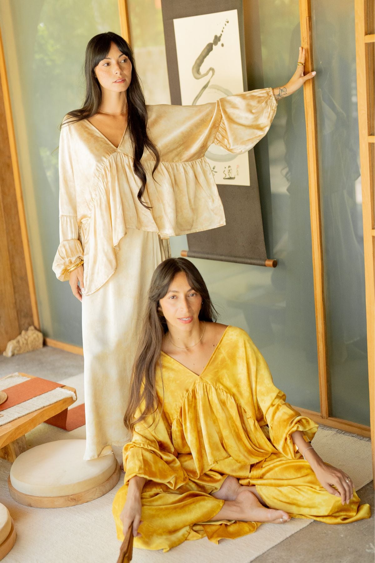 Two individuals are adorned in elegant ensembles made from 100% naturally dyed silk, courtesy of the Eva Top & Shanti Skirt Set by Myrah Penaloza. One wears a cream set while the other is in yellow. One stands gracefully touching a wall scroll, and the other sits on the floor. The setting features stylish wooden and glass accents.