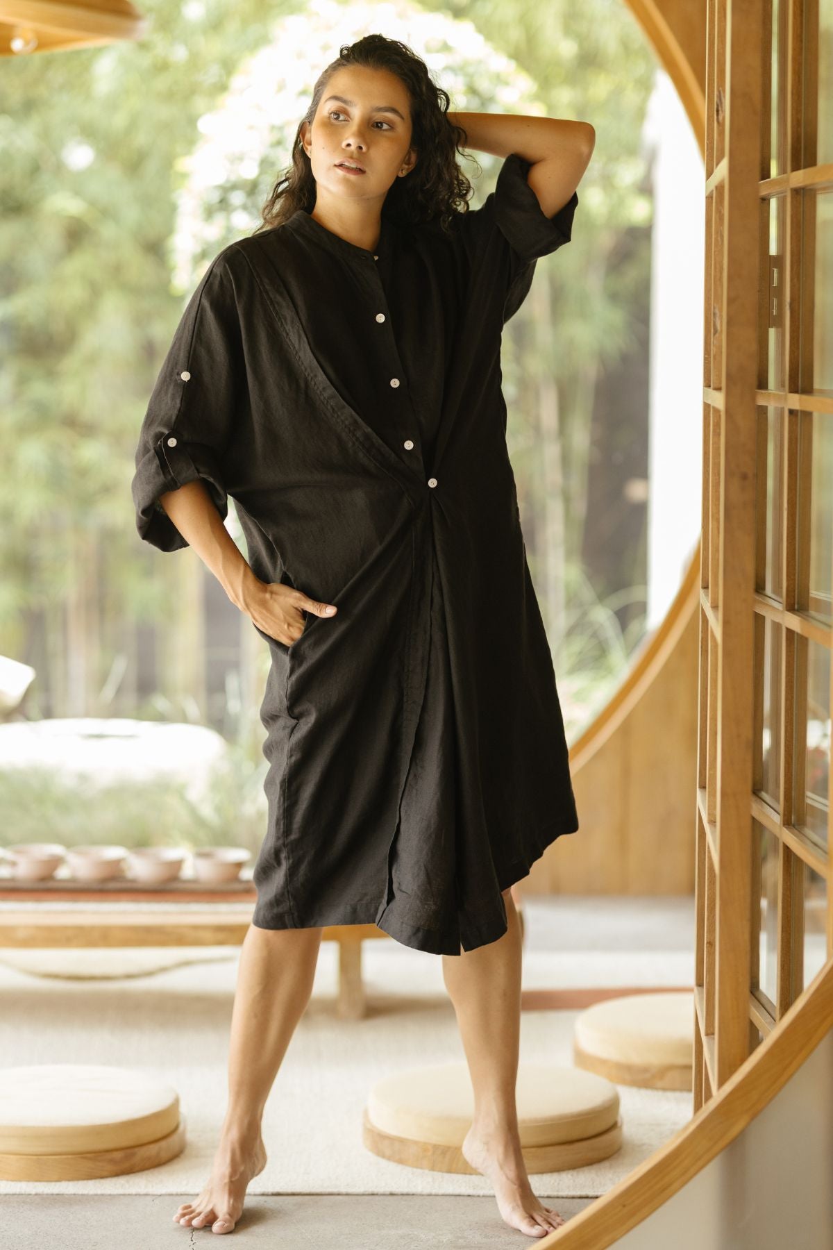 A woman stands barefoot indoors, wearing a loose-fitting black dress with buttons. One hand rests on her hip and the other on her head. She poses confidently in a well-lit room adorned with wooden furnishings and a round doorway in the background, showcasing the Myrah Penaloza Linen Still Water Tunic (Made To Order), crafted from 100% linen.