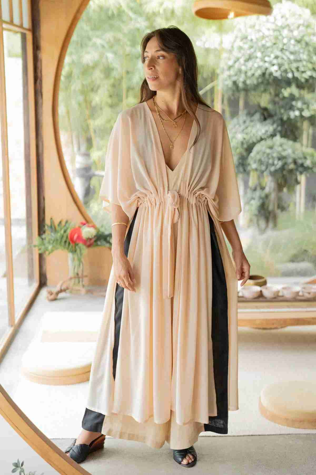 Indoors near a large round window with lush greenery, a woman stands wearing the Madelin Sheer Chiffon Long Cover by Myrah Penaloza over black pants and black sandals. The flowy, peach-colored fabric of the cover exudes sheer brilliance. Her long, dark hair cascades loosely as she rests one hand on her hip and gazes to the side.