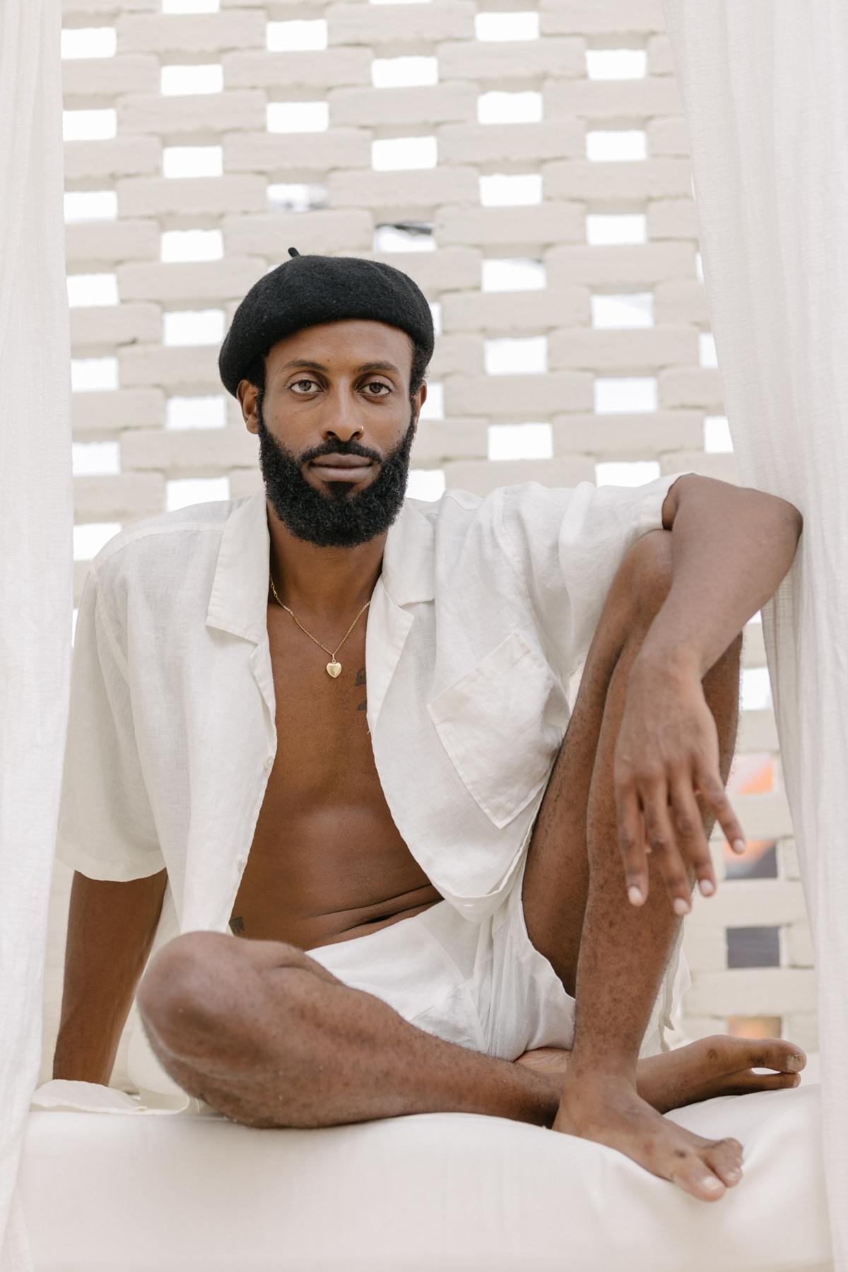 A nomadic traveler sits cross-legged on a white cushioned surface, wearing the Caravana Men's Linen Top & Short by Myrah Penaloza and a black beret. The sunlight filters gently through the white brick wall, enhancing the relaxed atmosphere with its lightweight and breathable charm.