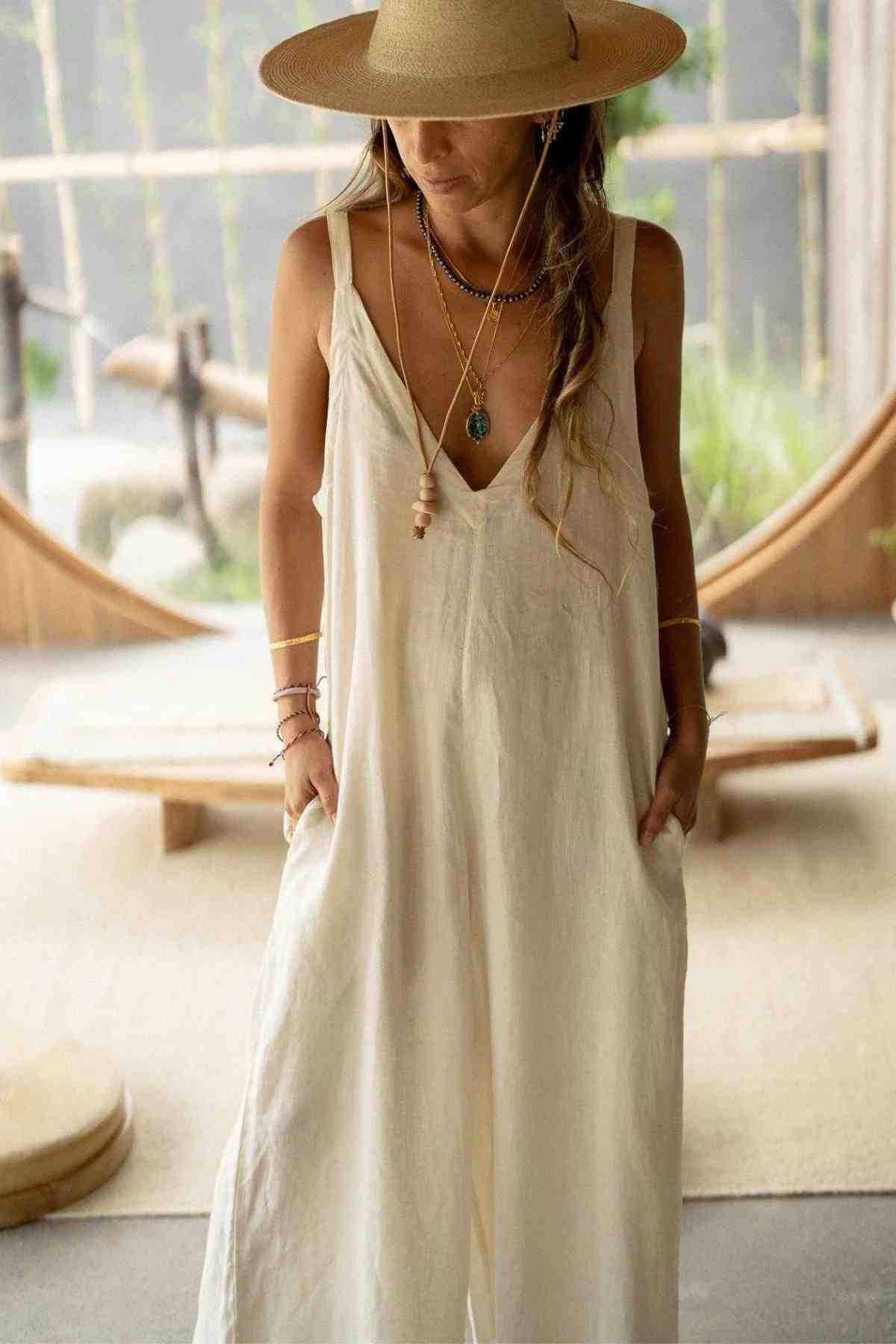 A woman stands indoors wearing the Kundalini Linen Playsuit by Myrah Penaloza, showcasing its luxuriously soft, stonewashed linen material and practical pockets. She accessorizes with layered necklaces and bracelets. Her long, wavy hair is partially tucked under a wide-brimmed straw hat. The background features a minimalistic, serene setting.