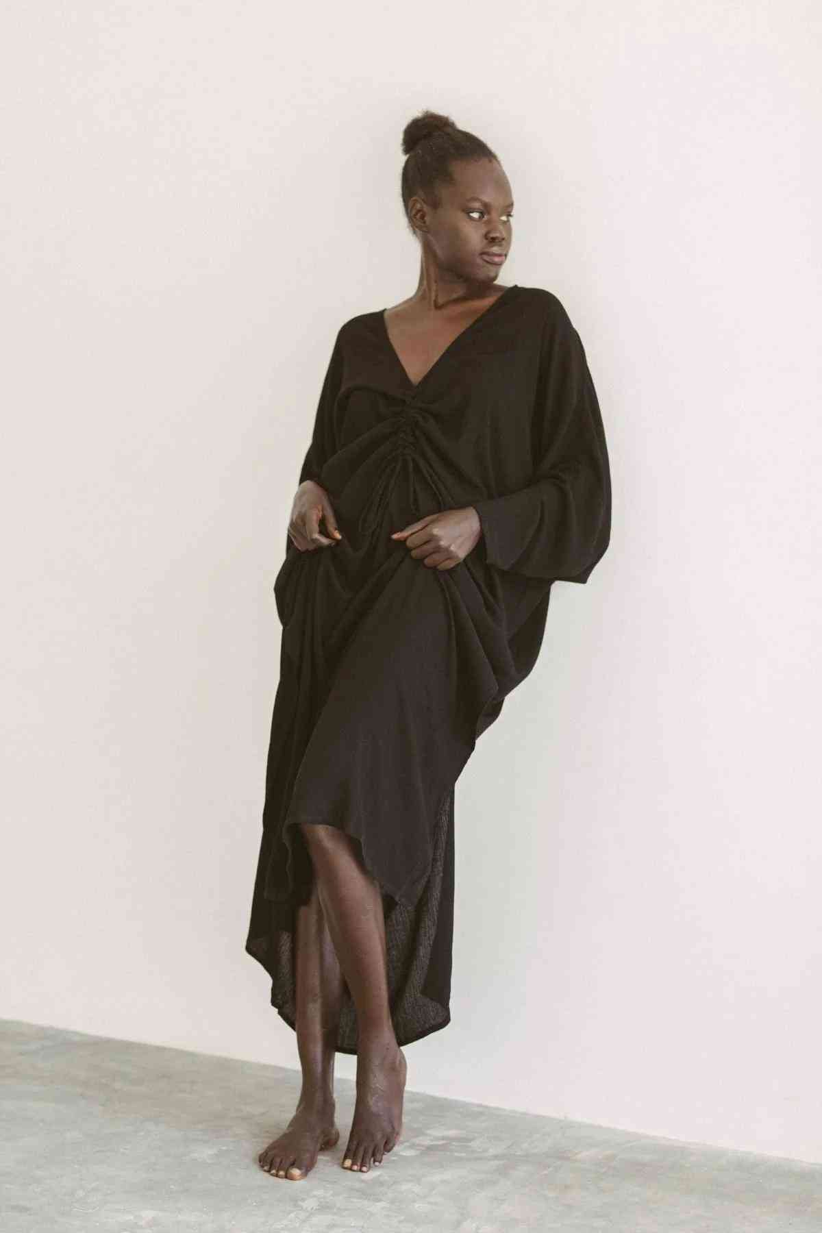 A person stands barefoot against a plain wall, wearing a loose-fitting, black Bamboo Rayon Virgo Rising Gown with long sleeves and a knot detail at the front. Their hair is styled in a bun, and they have one hand in a pocket while gazing to the side. The gown is from the brand Myrah Penaloza.