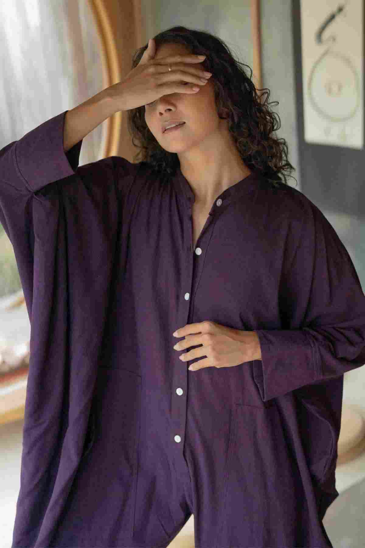 A person with long curly hair, wearing a loose-fitting, deep purple Miracle Playsuit 2.0 from Myrah Penaloza's new Karama Collection, stands with one hand covering their eyes. They seem relaxed, and soft natural light fills the room from a nearby window. A picture with abstract art is visible in the background.