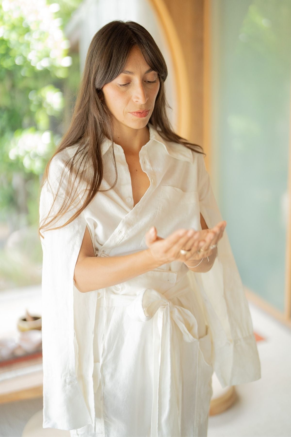A person with long dark hair stands in a brightly lit indoor space, gazing down at their open hands. Clad in the versatile Myrah Penaloza Sutra Linen Set, they wear a white, long-sleeved outfit that ties at the waist. Sunlight streams through the windows, illuminating green foliage outside.