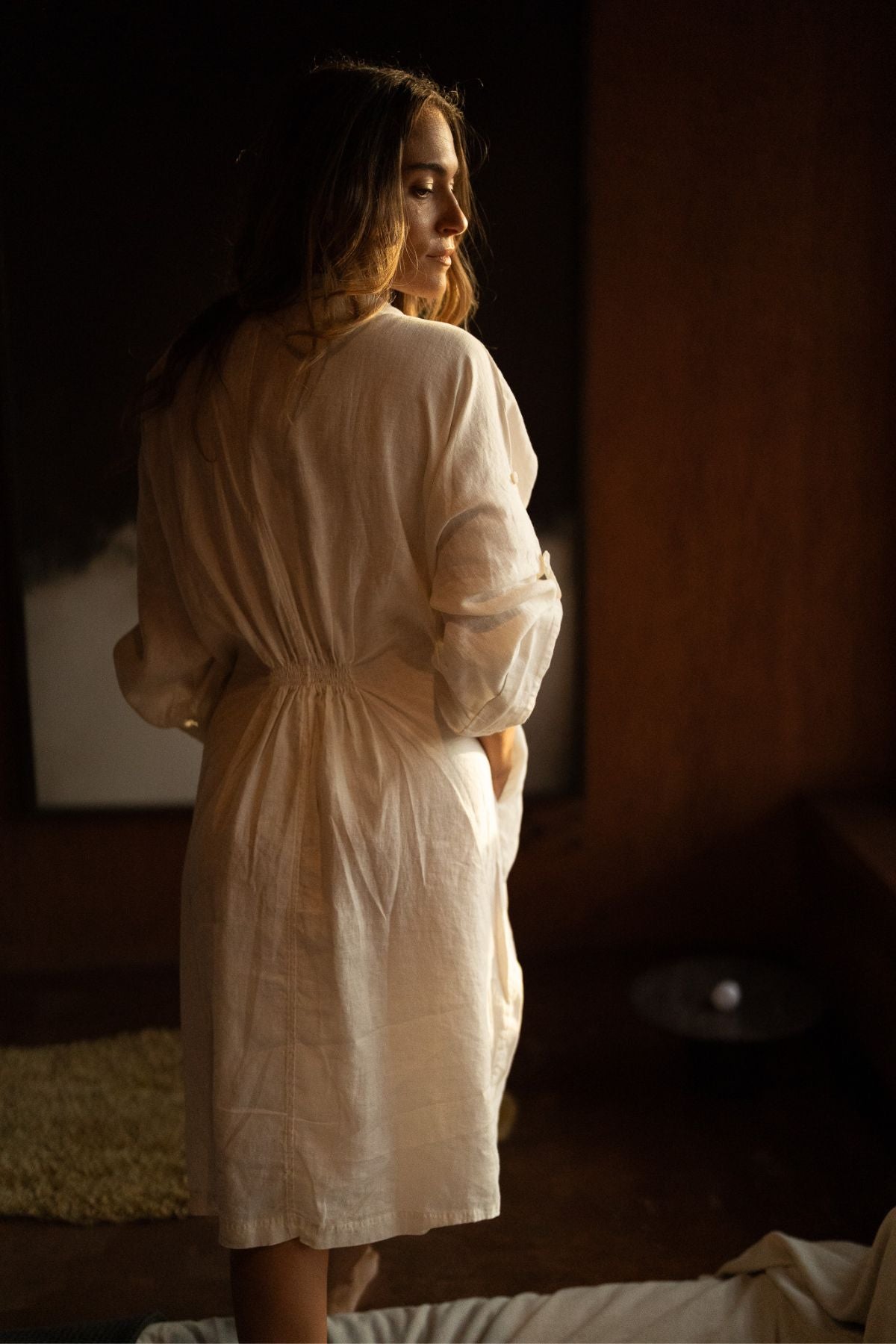 A woman wearing a light-colored, 100% linen Still Water Tunic from Myrah Penaloza's Made To Order collection stands in a dimly lit room. She faces away from the camera, turning her head to the side as her long hair cascades over her shoulders. The room features earthy tones and minimalistic decor.