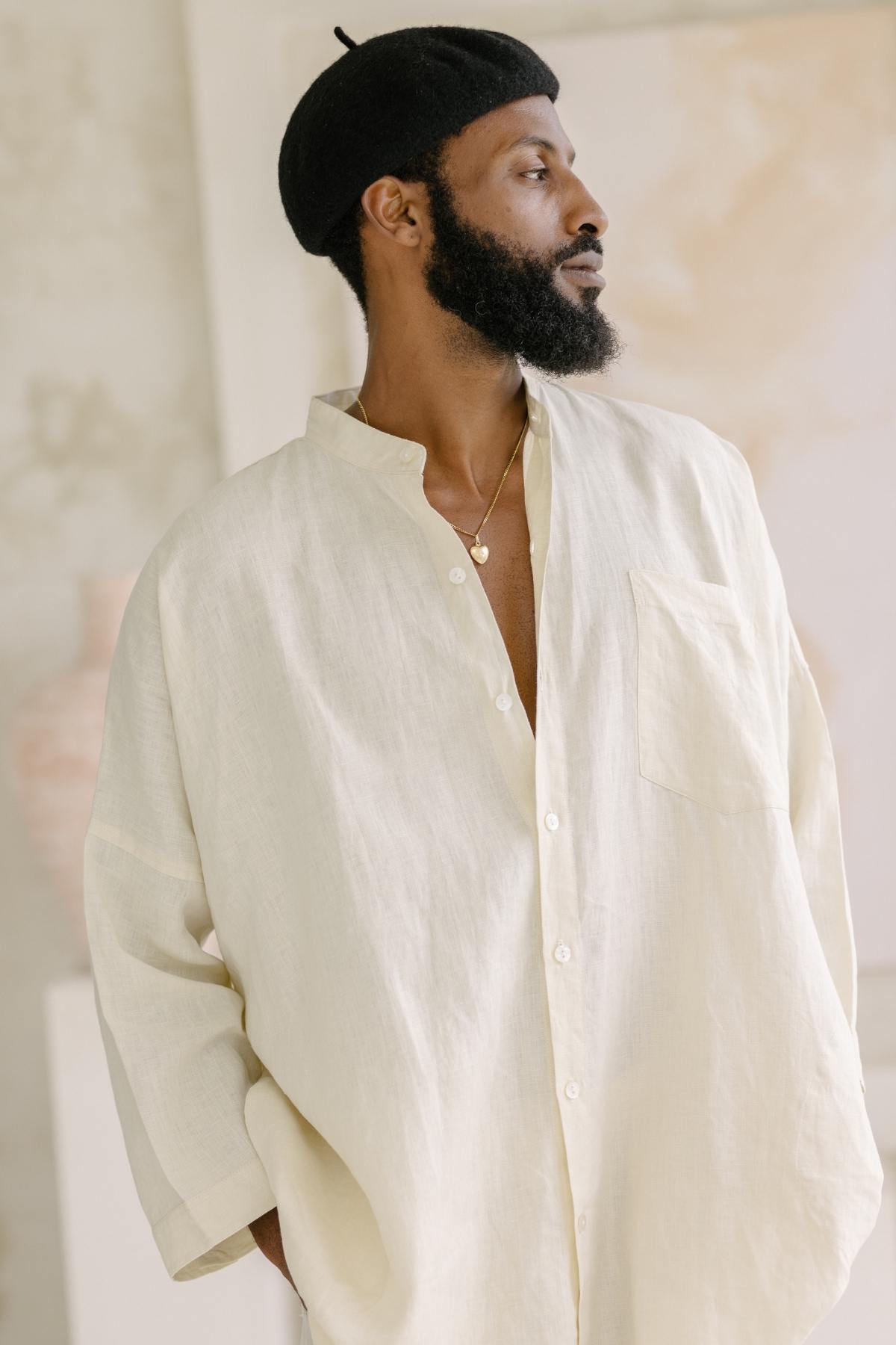 A person wearing a Camisa del Novio Mens Linen Top by Myrah Penaloza and a black beret looks to the side. They have a beard and are adorned with a gold necklace. The softly lit background features a blurred vase and artwork, adding an artistic touch.