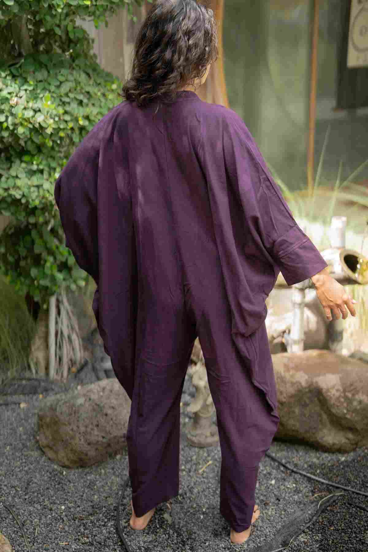 A person with long hair is standing outdoors with their back to the camera, wearing a loose-fitting, dark purple Miracle Playsuit 2.0 from the Karama Collection by Myrah Penaloza. The ground is covered with pebbles, and there are large rocks and lush greenery in the background.
