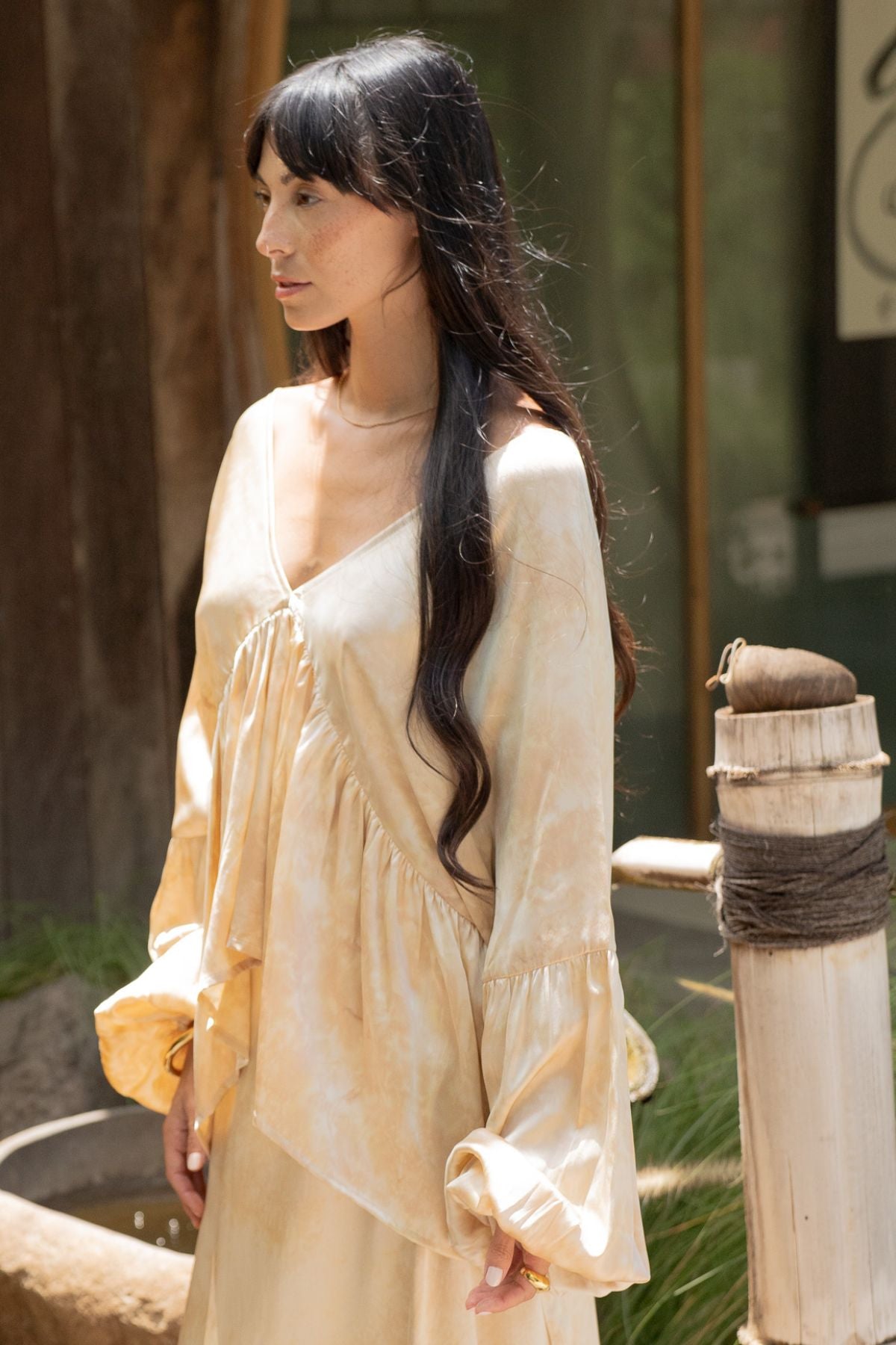 A woman with long dark hair stands outside wearing a light beige, flowing top with bell sleeves from the Myrah Penaloza Eva Top & Shanti Skirt Set, crafted from 100% naturally dyed silk. She is looking to the side, and the background includes a wooden post and some greenery.