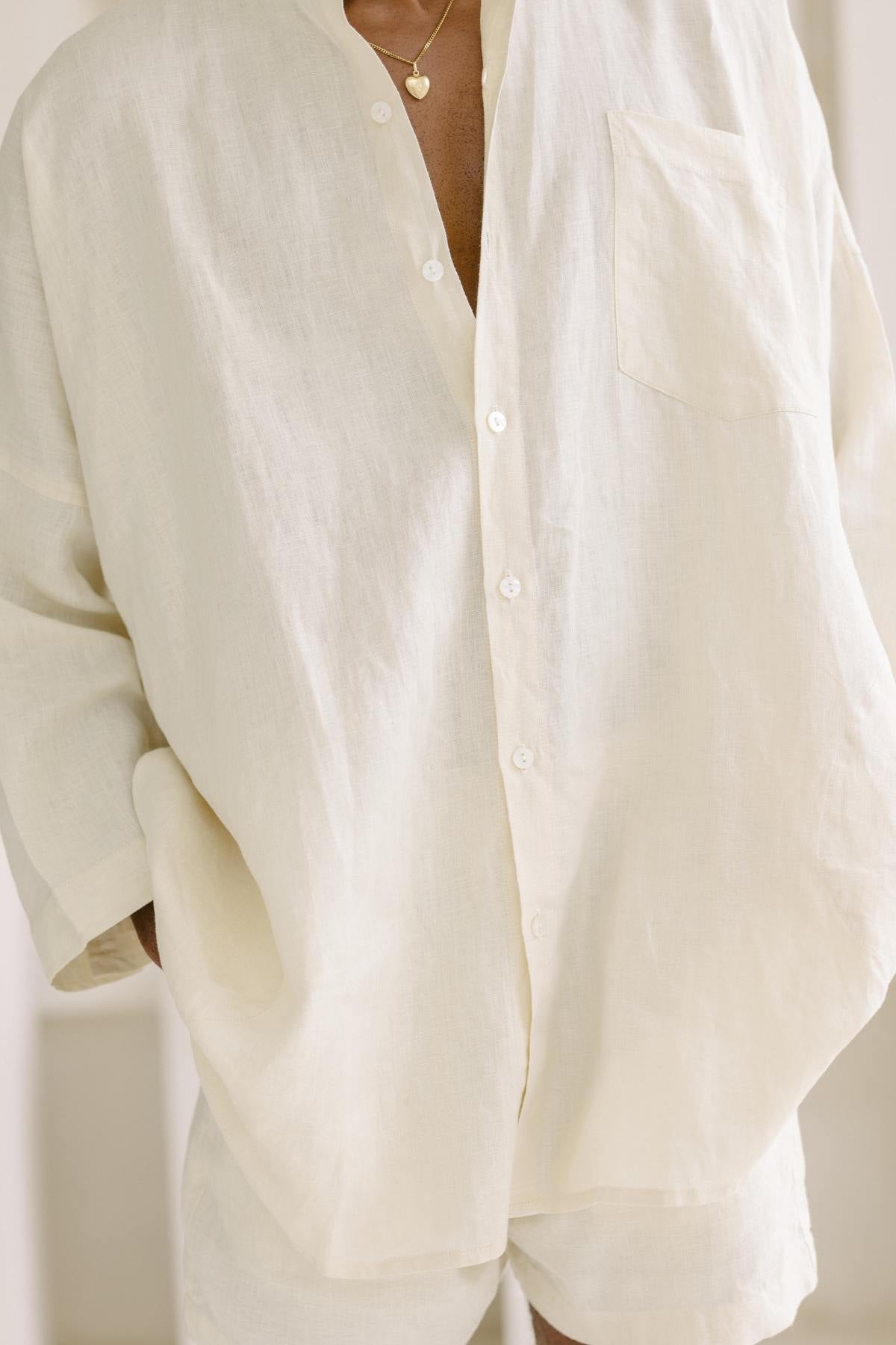 A person is dressed in the Camisa del Novio Mens Linen Top by Myrah Penaloza, a loose-fitting, button-up beige shirt with a classic boyfriend style, paired with shorts. A small gold necklace gleams subtly against the light and comfortable stone-washed linen fabric. The background features an elegantly blurred effect.