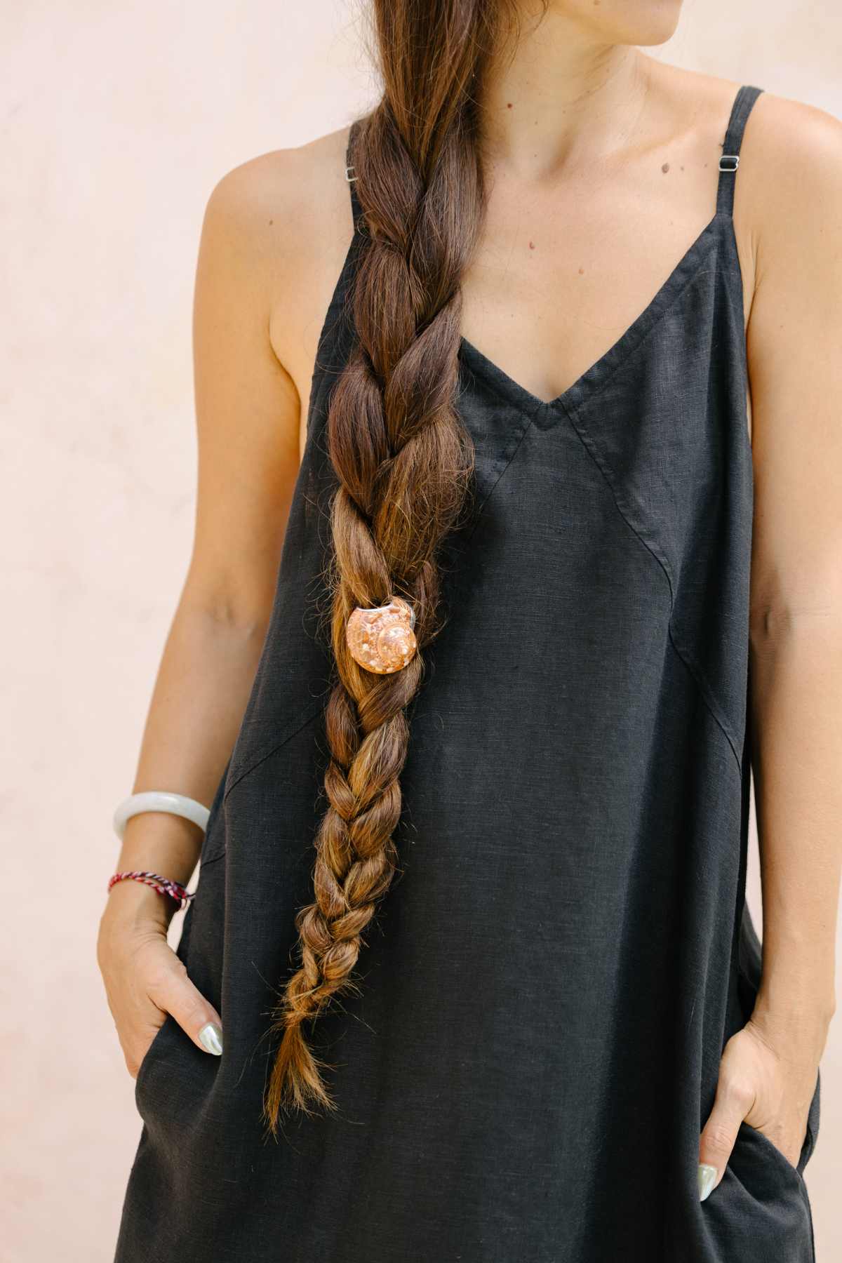 A person with a long, thick braid adorned with a decorative hair tie and extending over one shoulder is pictured from the chest down, wearing an Everyday Linen Slip by Myrah Penaloza. The sleeveless black dress flows elegantly as their hands rest casually in their pockets, creating a relaxed yet sophisticated look.