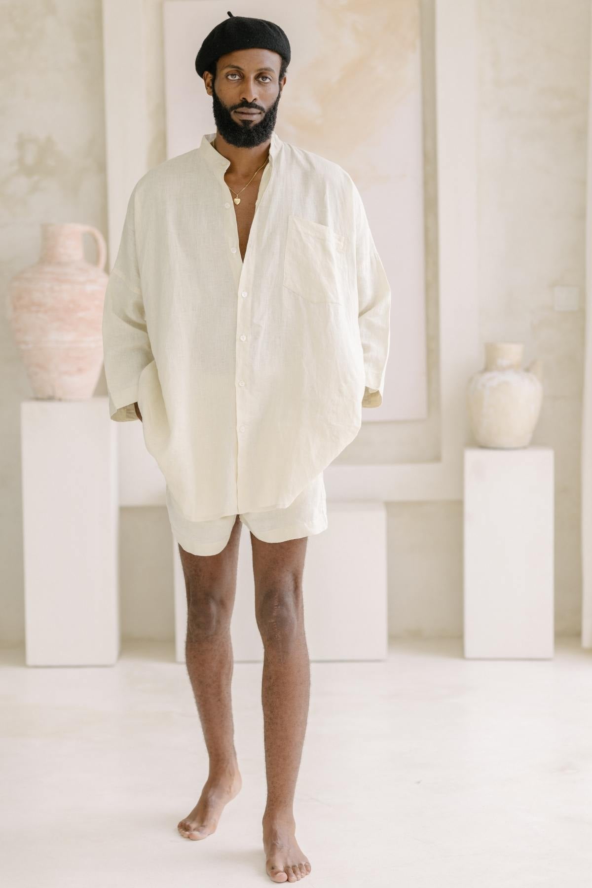 A person with a beard and a beret is wearing an oversized light beige Camisa del Novio Mens Linen Top by Myrah Penaloza, paired with matching shorts. They are standing barefoot in a minimalist room, where stone-washed linen textures complement the neutral-colored pottery displayed on pedestals in the background.