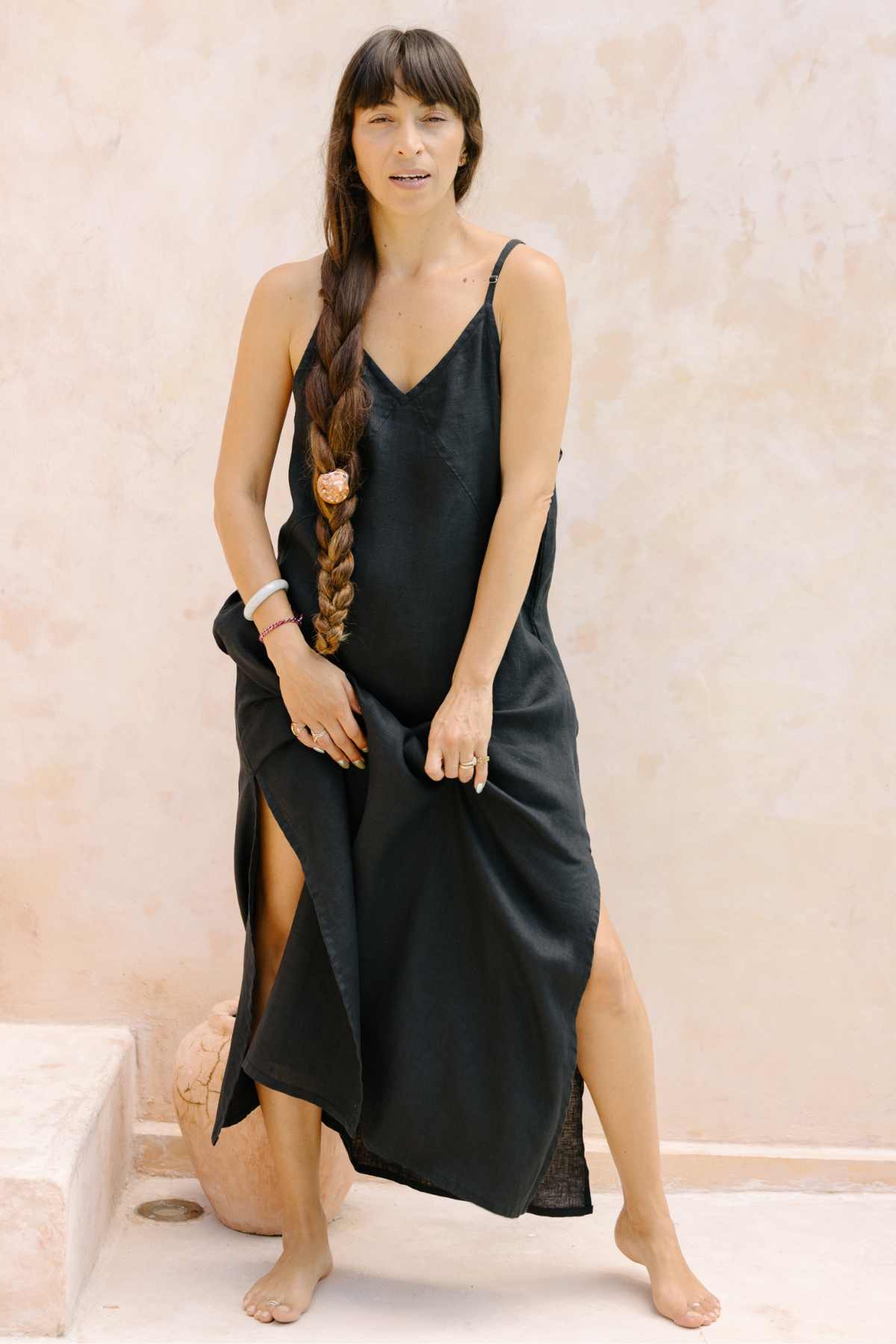 A woman stands barefoot against a light-colored wall, adorned in the sleeveless, long black Everyday Linen Slip by Myrah Penaloza, which features side slits. Her long, braided hair cascades over her shoulder. Embracing luxurious comfort, she holds the hem of the stone-washed linen slip with one hand and maintains a neutral expression.