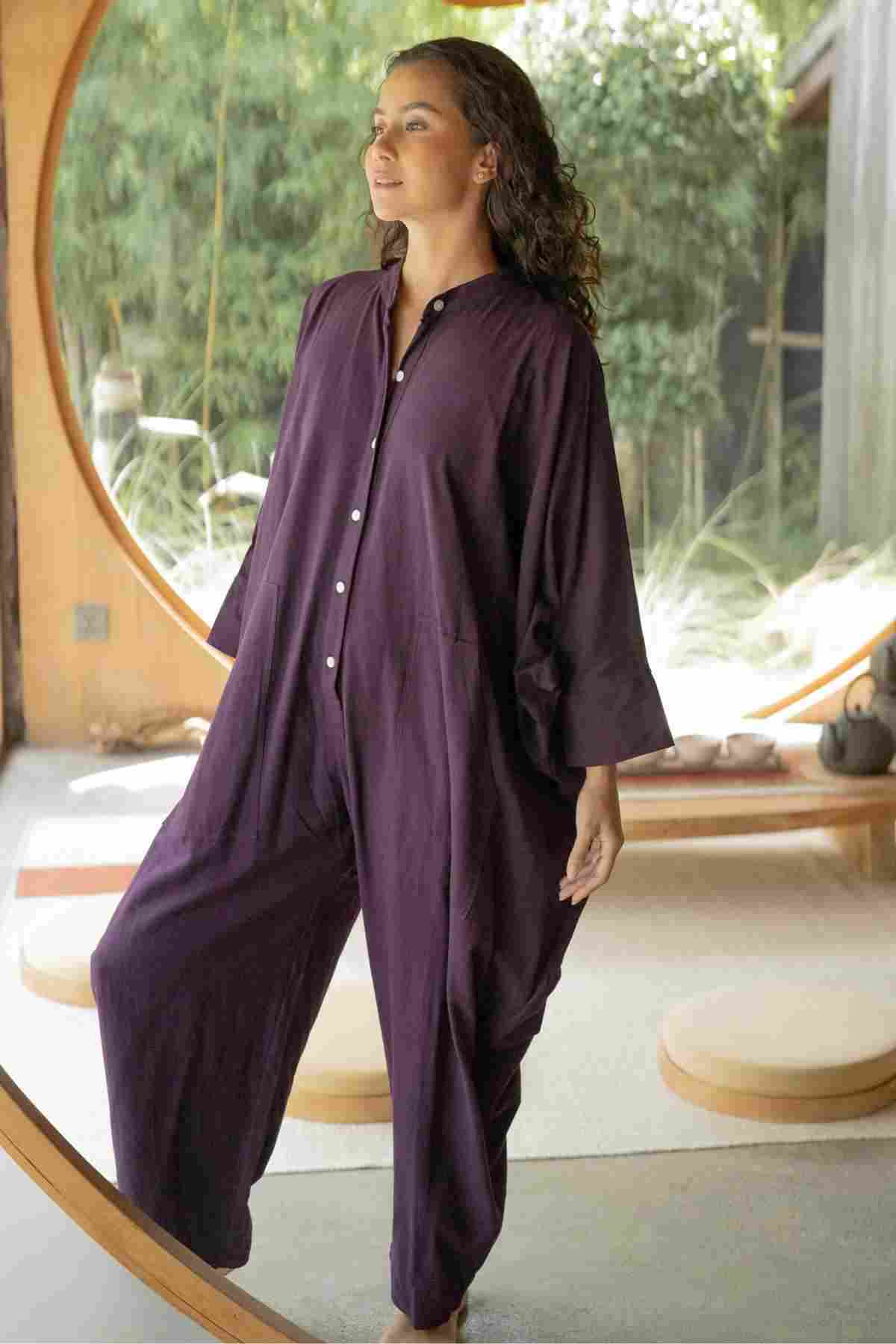 A person with curly hair stands inside a round doorway, wearing the limited edition Miracle Playsuit 2.0 from the Karama Collection in deep purple by Myrah Penaloza. They look off into the distance with a calm expression. The background features a wooden bench and green foliage outside.