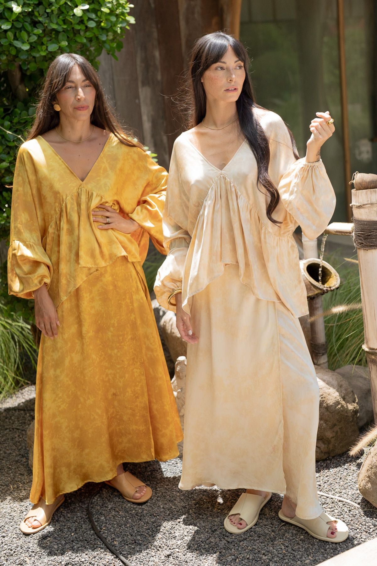 Two people stand outdoors, one wearing the golden yellow Shanti Skirt from the "Eva Top & Shanti Skirt Set" by Myrah Penaloza, crafted from 100% naturally dyed silk. The other is dressed in creamy beige sustainable linen. They are surrounded by greenery and rustic decor, creating a serene and natural setting.