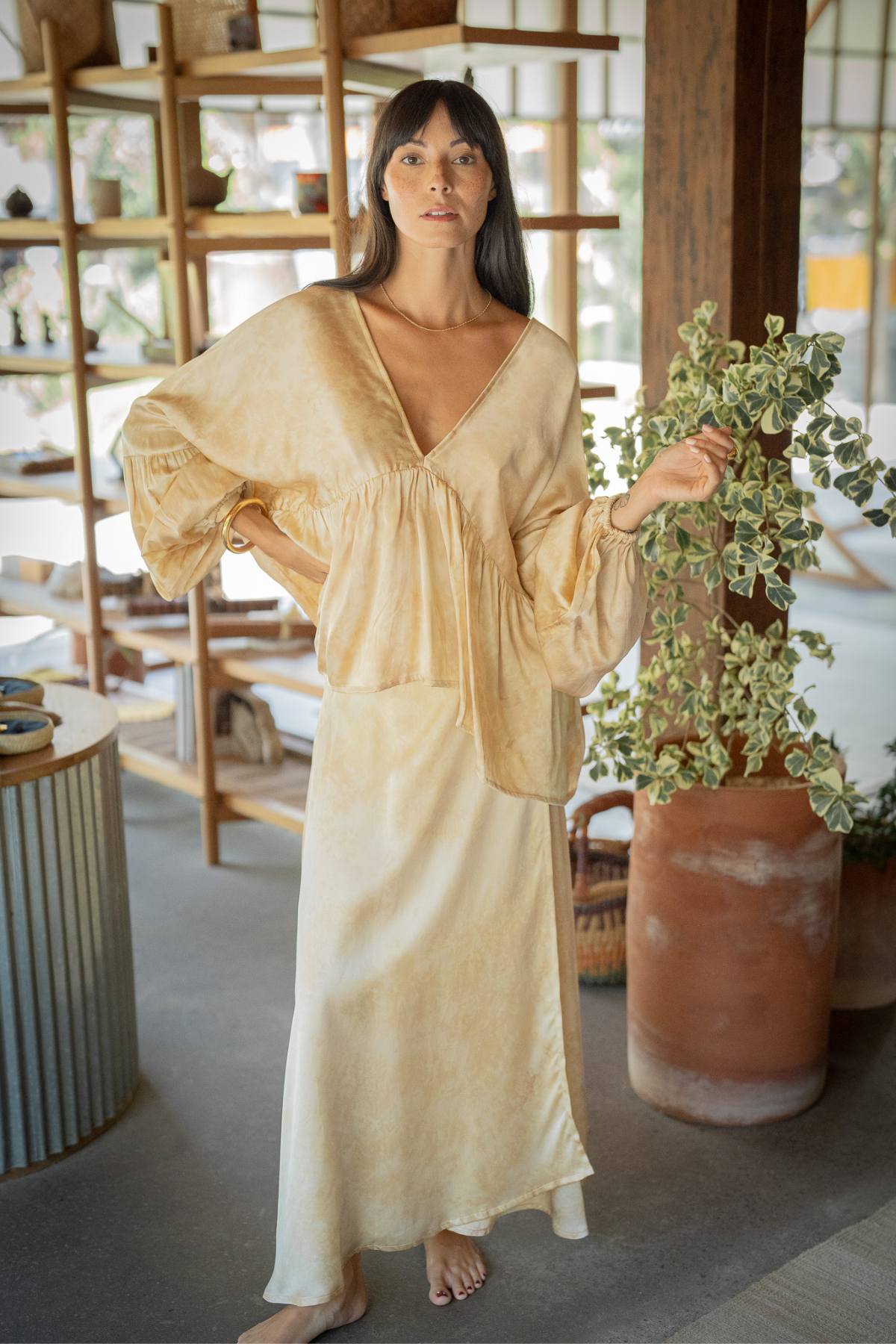 In a chic room adorned with wooden shelves and a leafy plant, a woman stands barefoot wearing the elegant Myrah Penaloza Eva Top & Shanti Skirt Set, made to order from 100% naturally dyed silk, as she gazes at the camera.