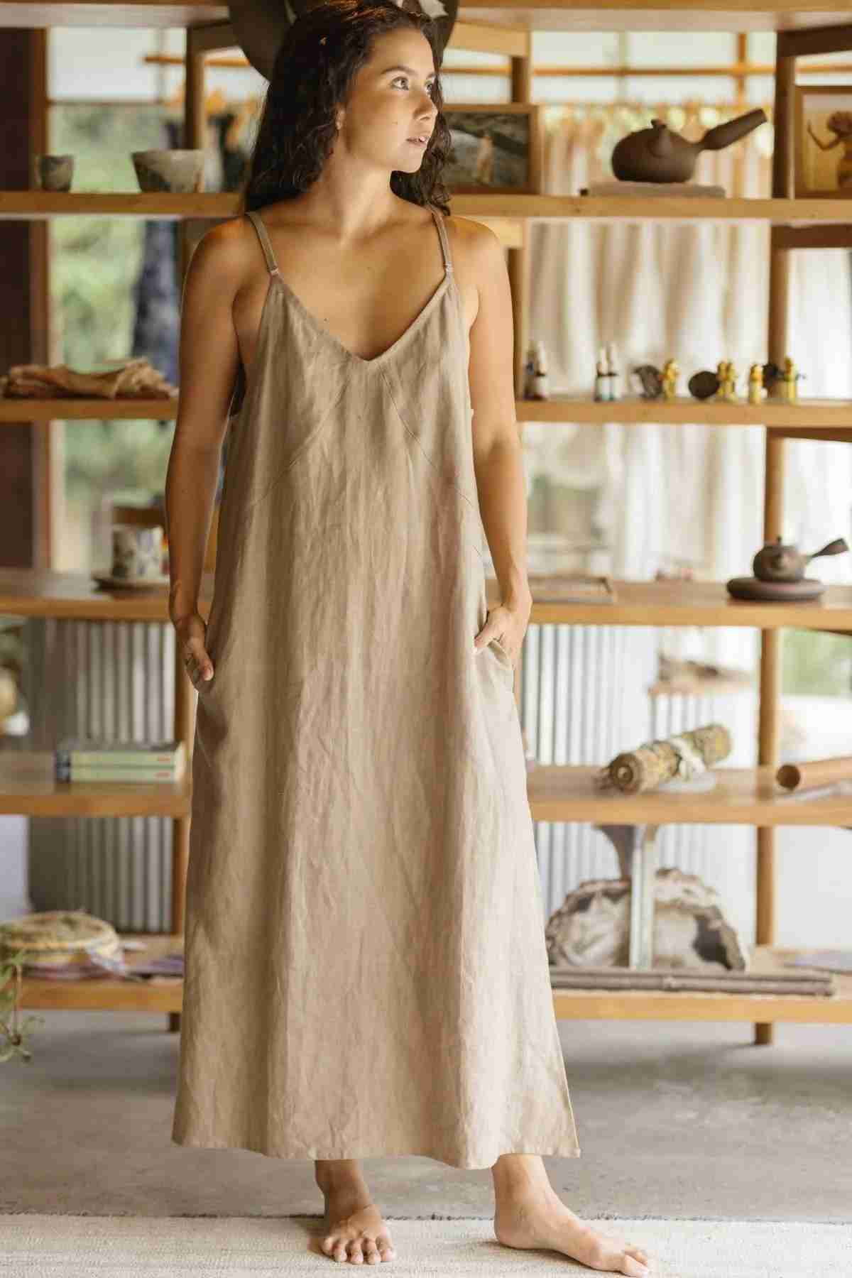 A woman stands barefoot in a softly lit, serene room adorned with wooden shelves displaying various items. Clad in an Everyday Linen Slip by Myrah Penaloza, she wears a long, sleeveless beige dress with thin straps made of stone-washed linen. Gazing to her right, she has her hands tucked into the pockets of her dress.