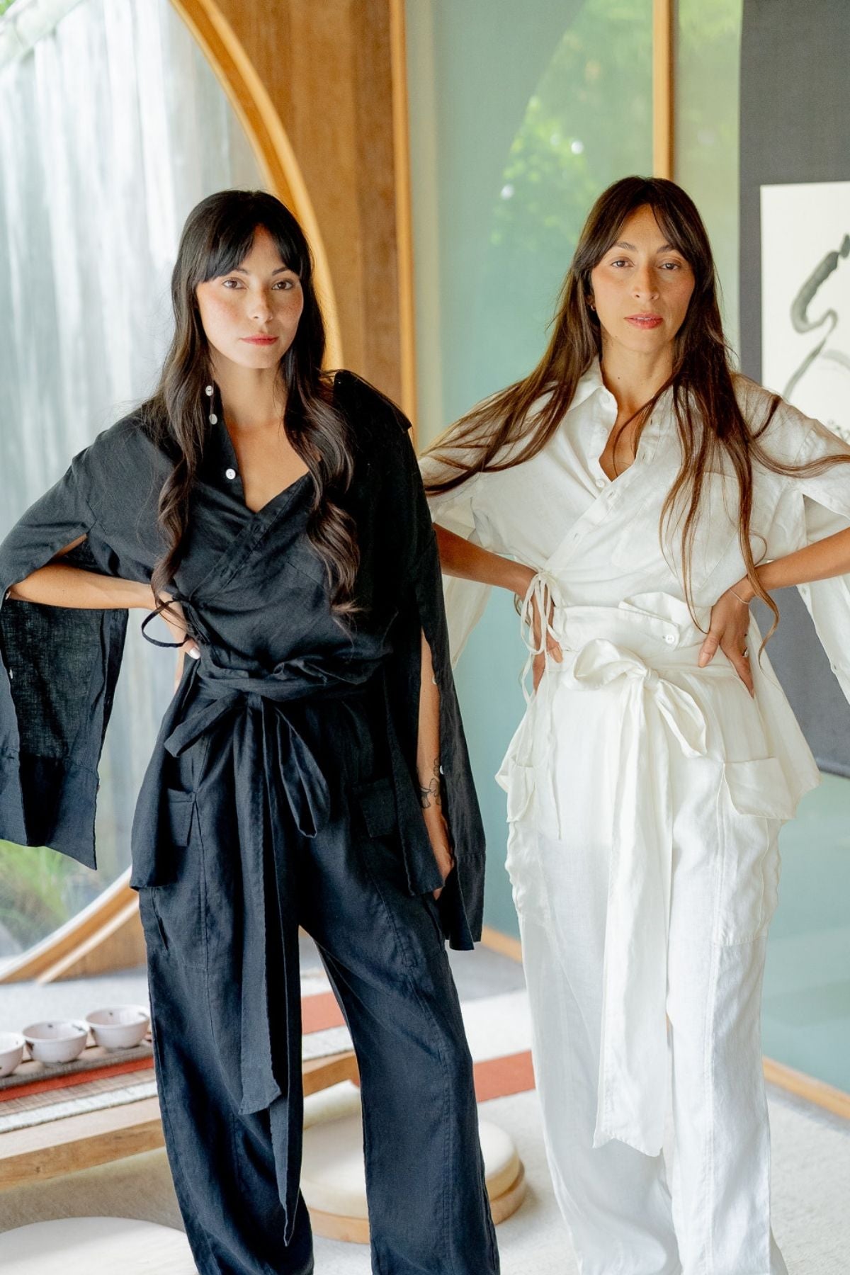 Two women, each with long dark hair, pose confidently indoors wearing minimalist, elegantly designed outfits. The woman on the left models a versatile black Sutra Linen Set from Myrah Penaloza's Made To Order collection, while the woman on the right sports a similar design in white lightweight linen. The room features a large circular window.