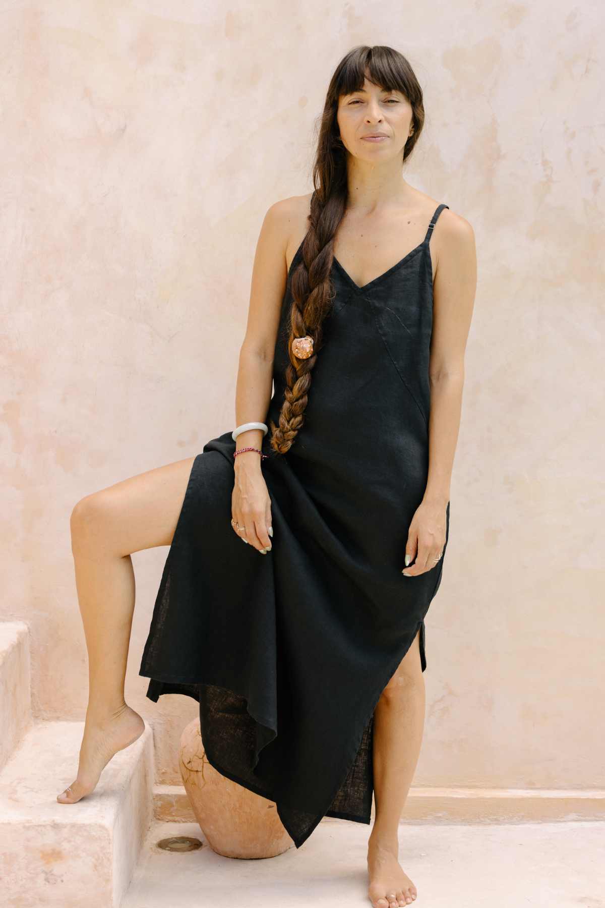 A woman with long, braided hair stands barefoot on a stone step indoors, wearing the Myrah Penaloza Everyday Linen Slip. Her neutral expression meets the camera's gaze. The sleeveless black dress exudes luxurious comfort against the light-colored, textured wall in the background.