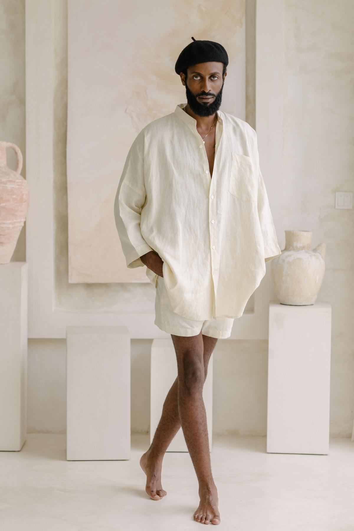 A person clad in the Myrah Penaloza Camisa del Novio Mens Linen Top and shorts stands barefoot in a neutral-toned room adorned with large vases and minimalistic decor. They wear a black beret and have a calm expression, perfectly complementing the serene stone-washed linen ambiance.